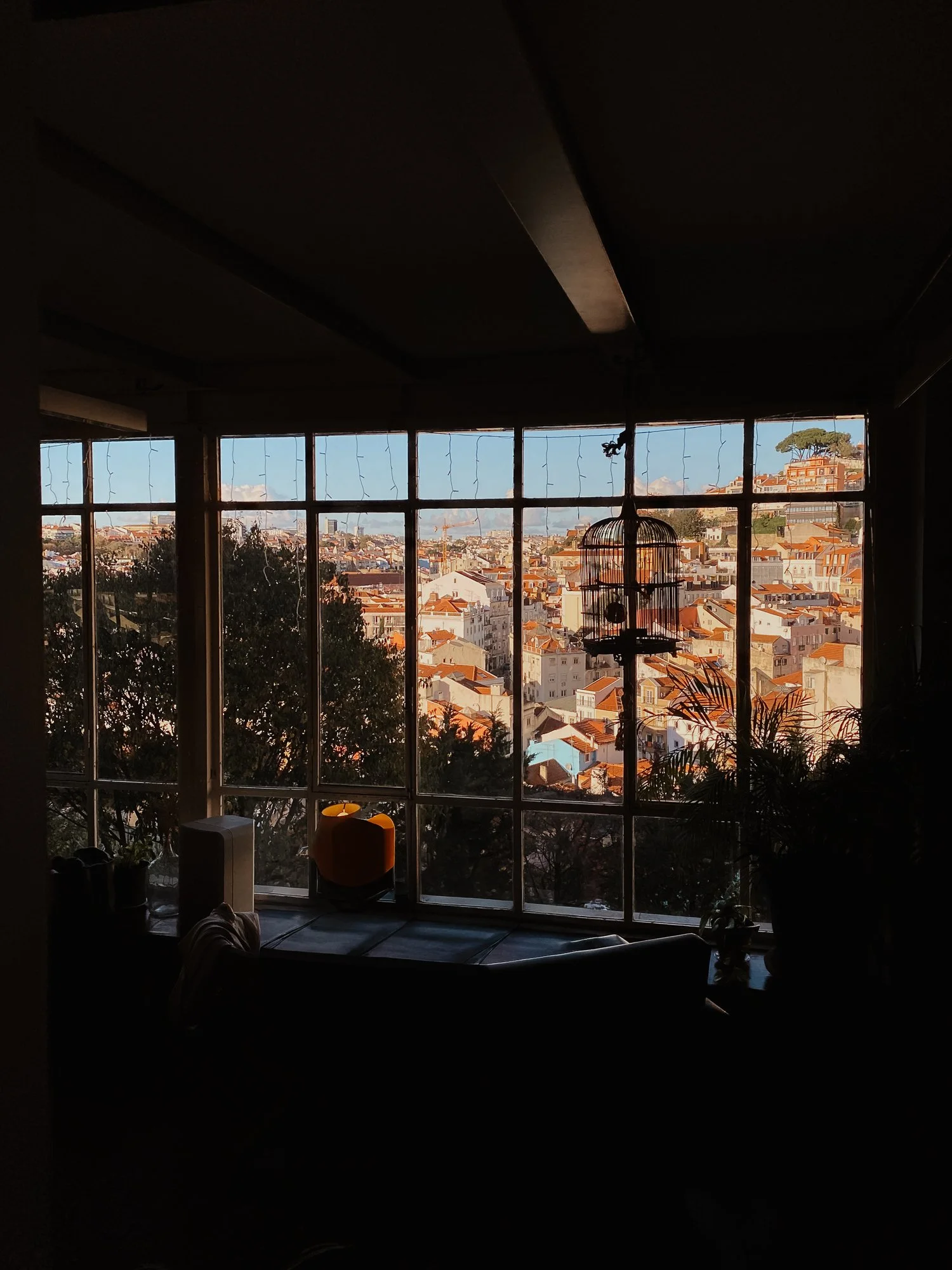 Innenraum mit Blick auf die Stadt durch große Fenster. Es hängen Lichterketten, ein Vogelkäfig und einige Pflanzen sind sichtbar.