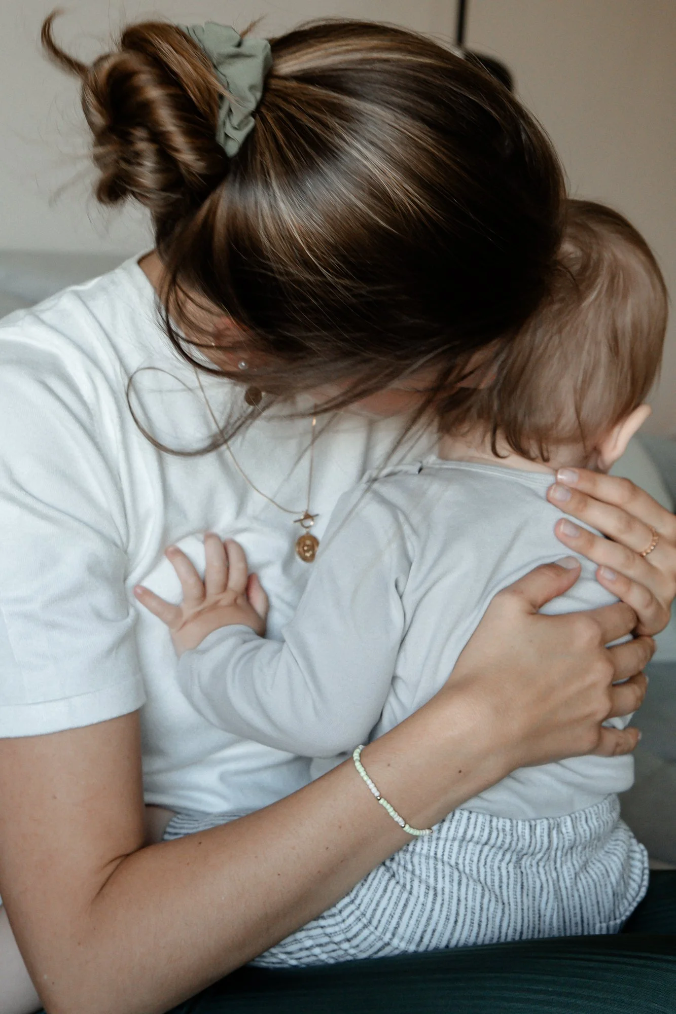 Eine Frau hält ein Kleinkind, das auf ihren Schultern sitzt, und gibt ihm einen Kuss. Die Frau trägt ein weißes T-Shirt, Halsketten und ein Armband.