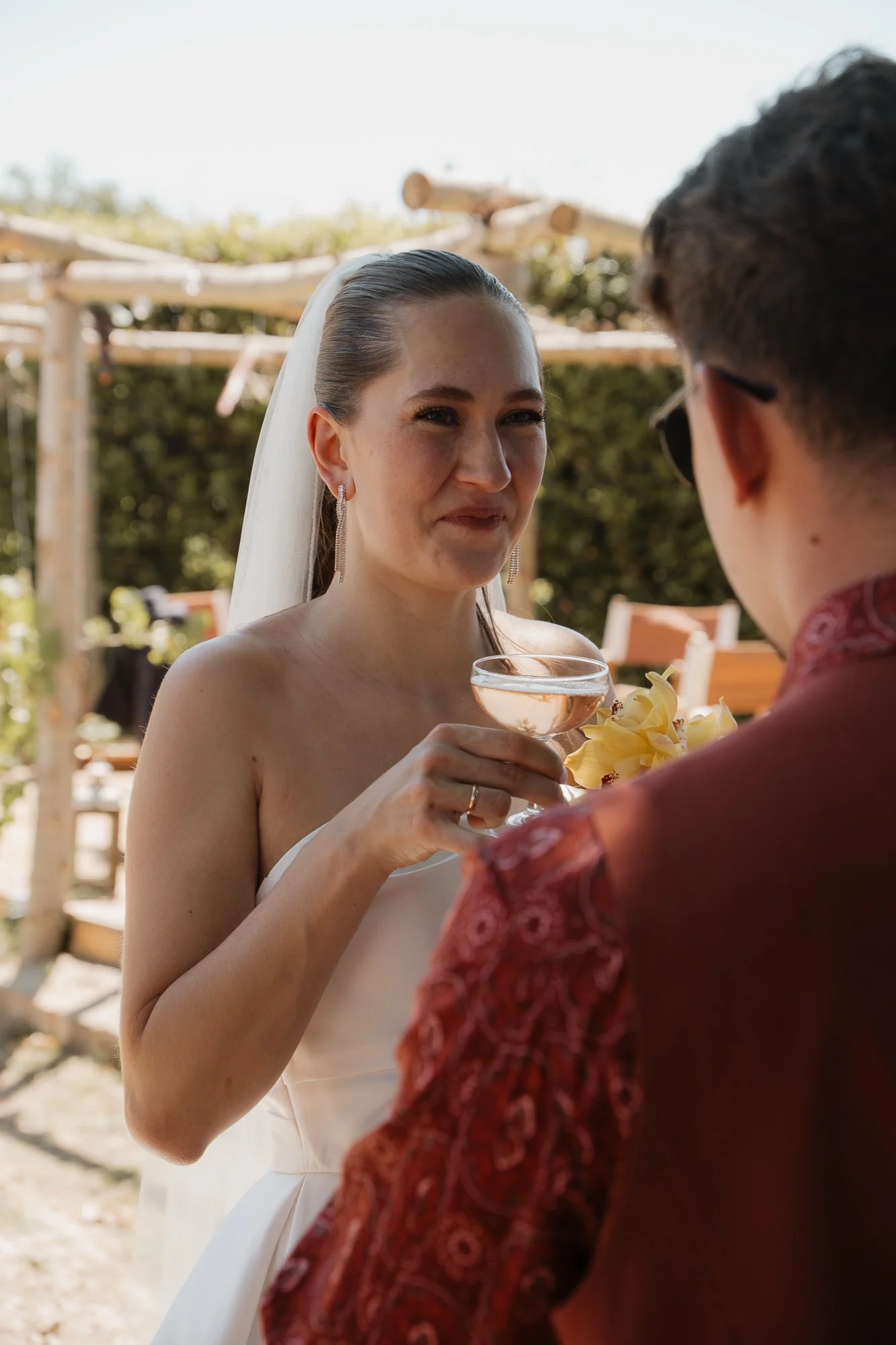 Eine Frau in einem weißen Hochzeitskleid hält ein Cocktailglas und trinkt bei einer Hochzeit im Freien, im Hintergrund sind Bäume und Holzbalken zu sehen.