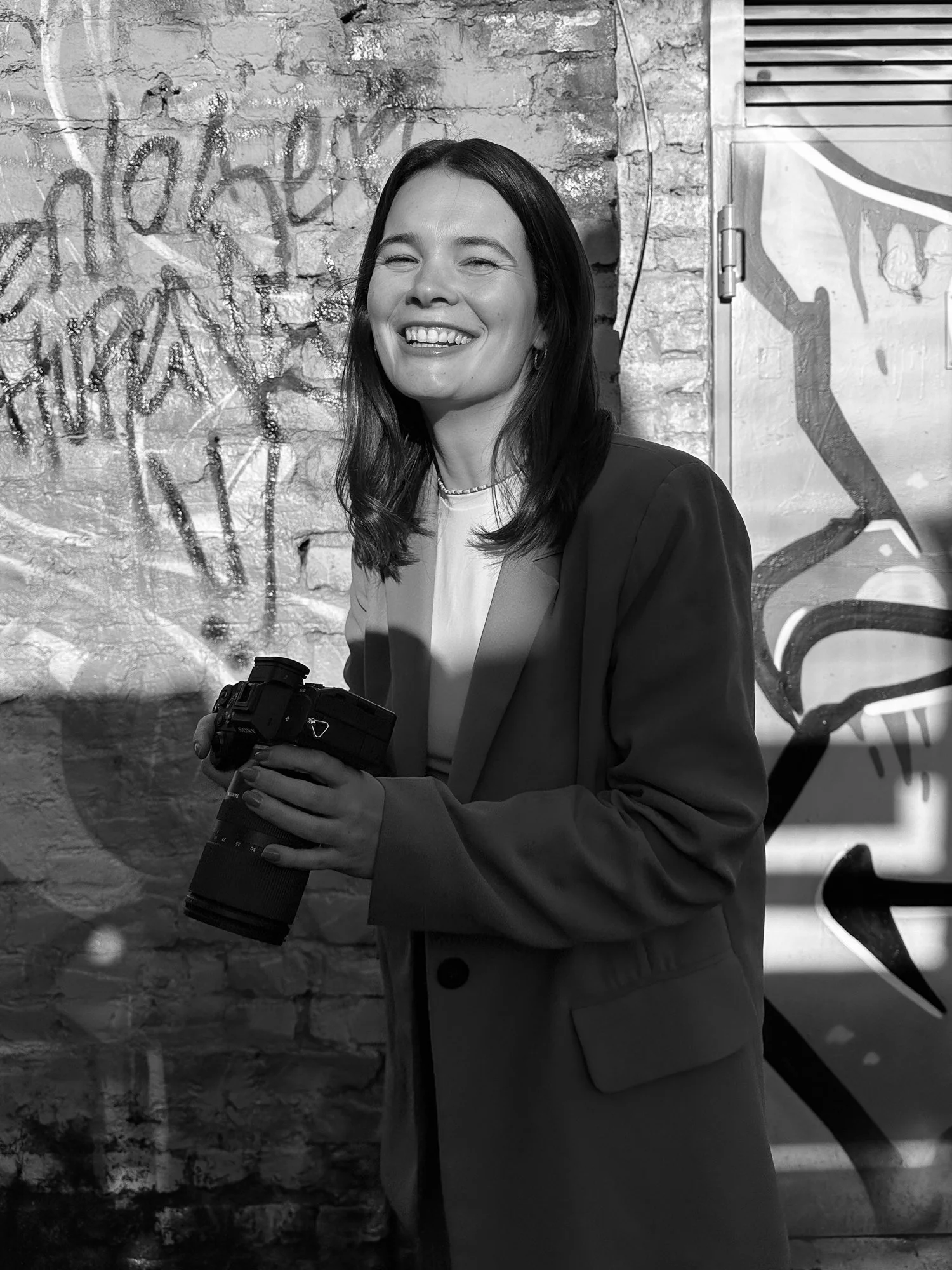 Junge Frau lächelt lachend in Schwarzweiß, hält eine Kamera, steht vor einer Graffiti-Wand aus Ziegelsteinen.