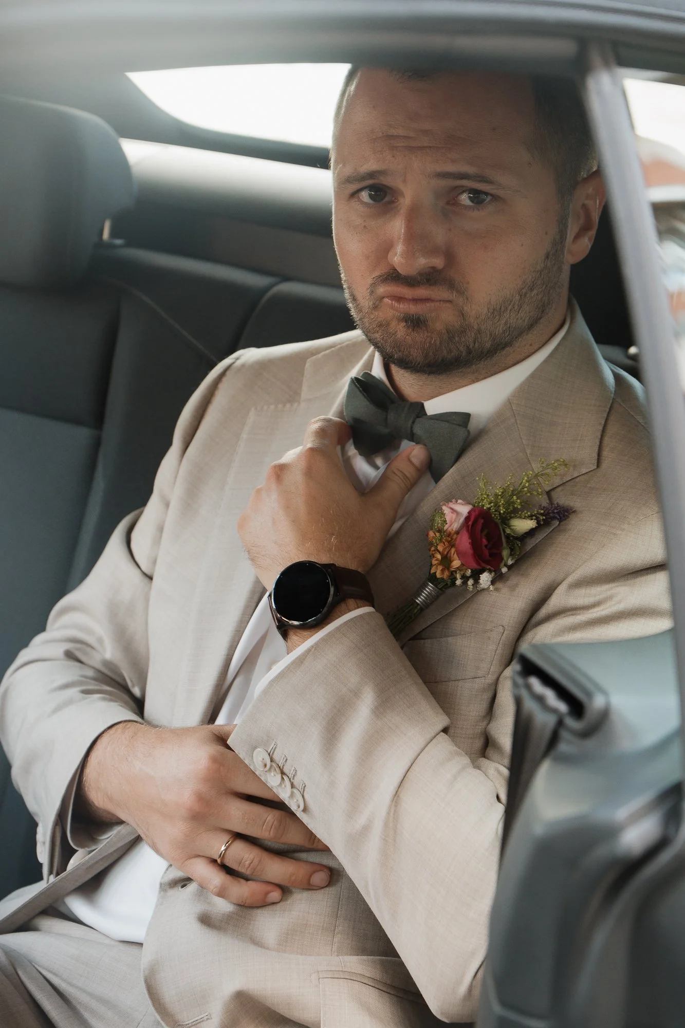 Bräutigam in Anzug mit Fliege und Blumenkrawatte, sitzt im Auto und schaut ernst.