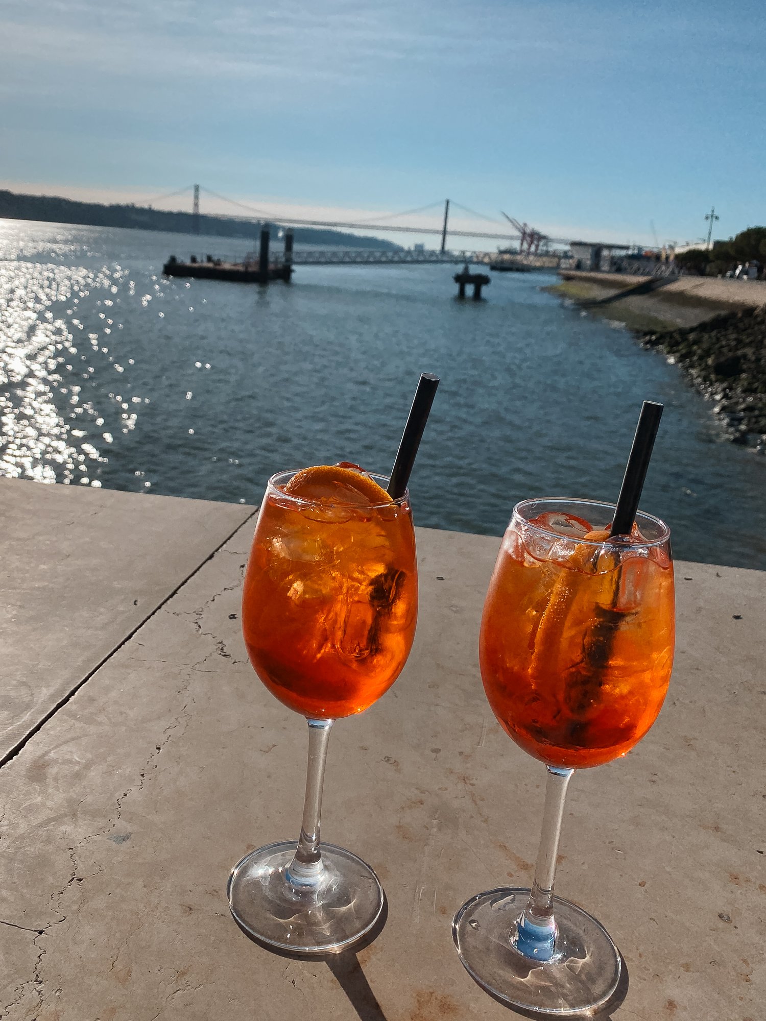 Zwei Gläser mit Aperol Spritz am Hafen mit Blick auf das Wasser, eine Brücke und das Ufer bei Sonnenlicht.