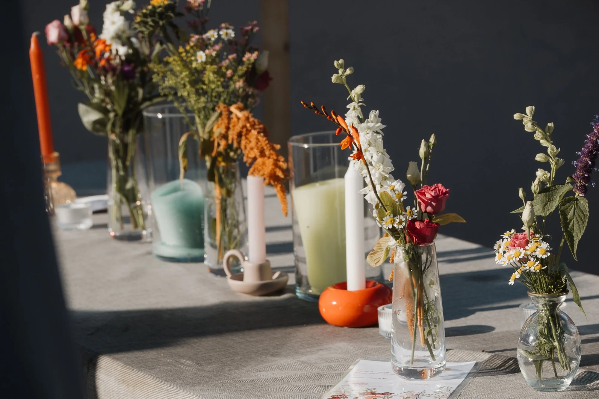 Blumenarrangement mit bunten Blumen und Kerzen auf einem Tisch bei Tageslicht.