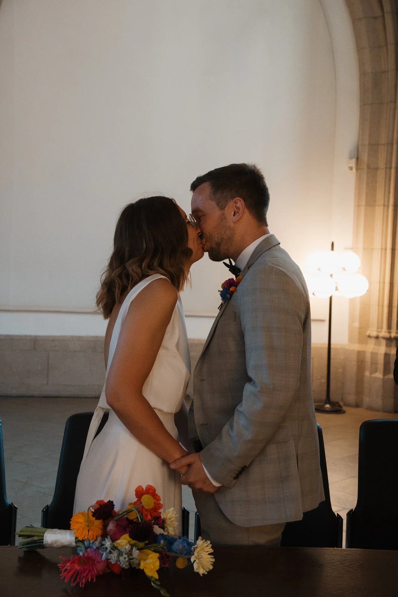 Ein Brautpaar bei einer Hochzeit, das sich küsst und Händchen hält, im Hintergrund eine Lampe und eine große leere Wand.