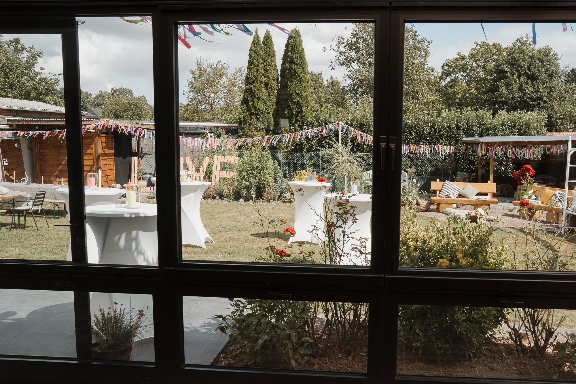 Gartenparty im Freien mit festlich dekorierten Tischen und Sitzgelegenheiten, buntes Girlanden-Dekor, und grünen Bäumen im Hintergrund.