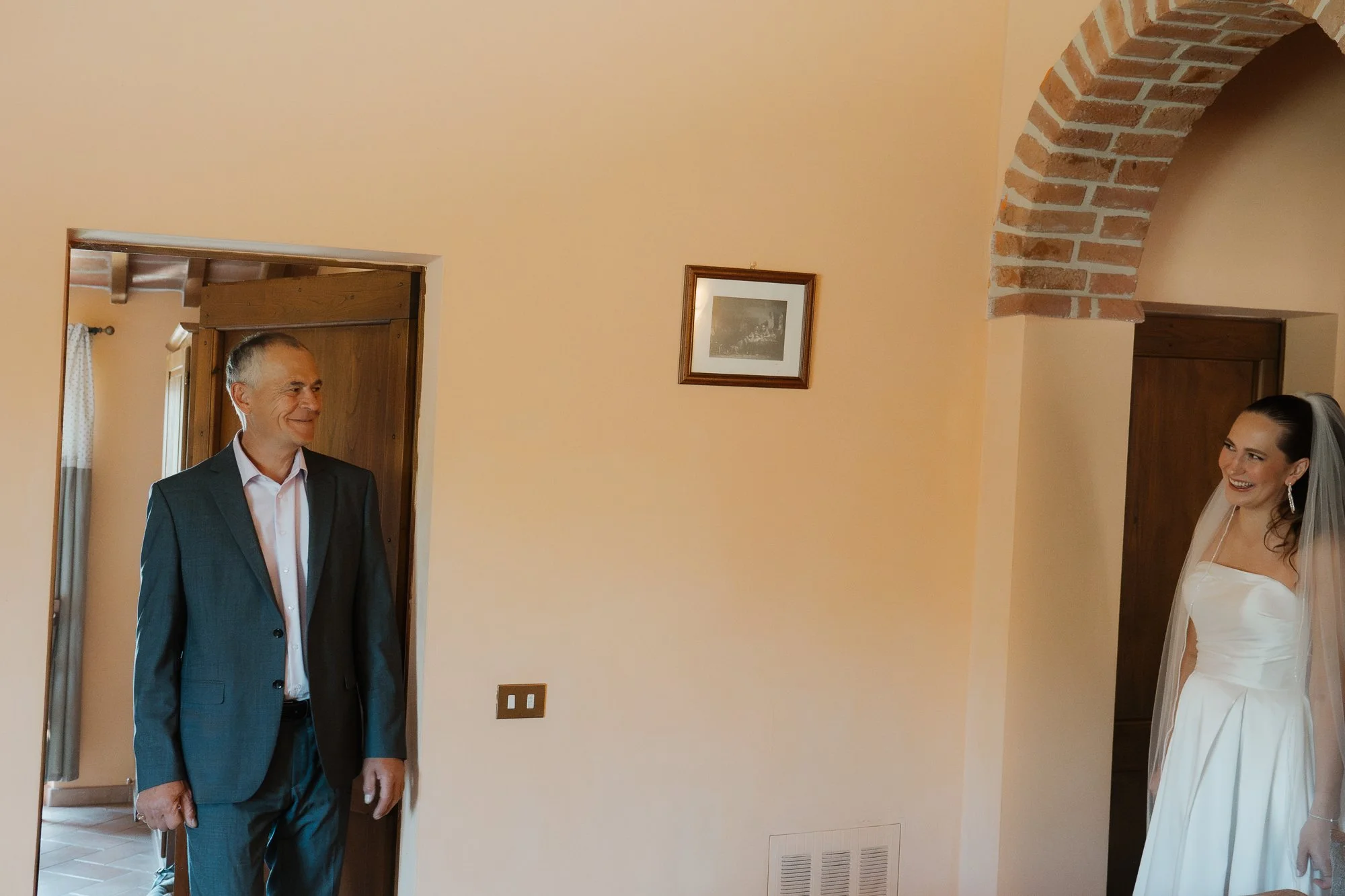 Ein Mann in Anzug und eine braut in Brautkleid und veil stehen sich gegenüber in einem Raum mit hellen Wänden und einem kleinen Bild an der Wand.