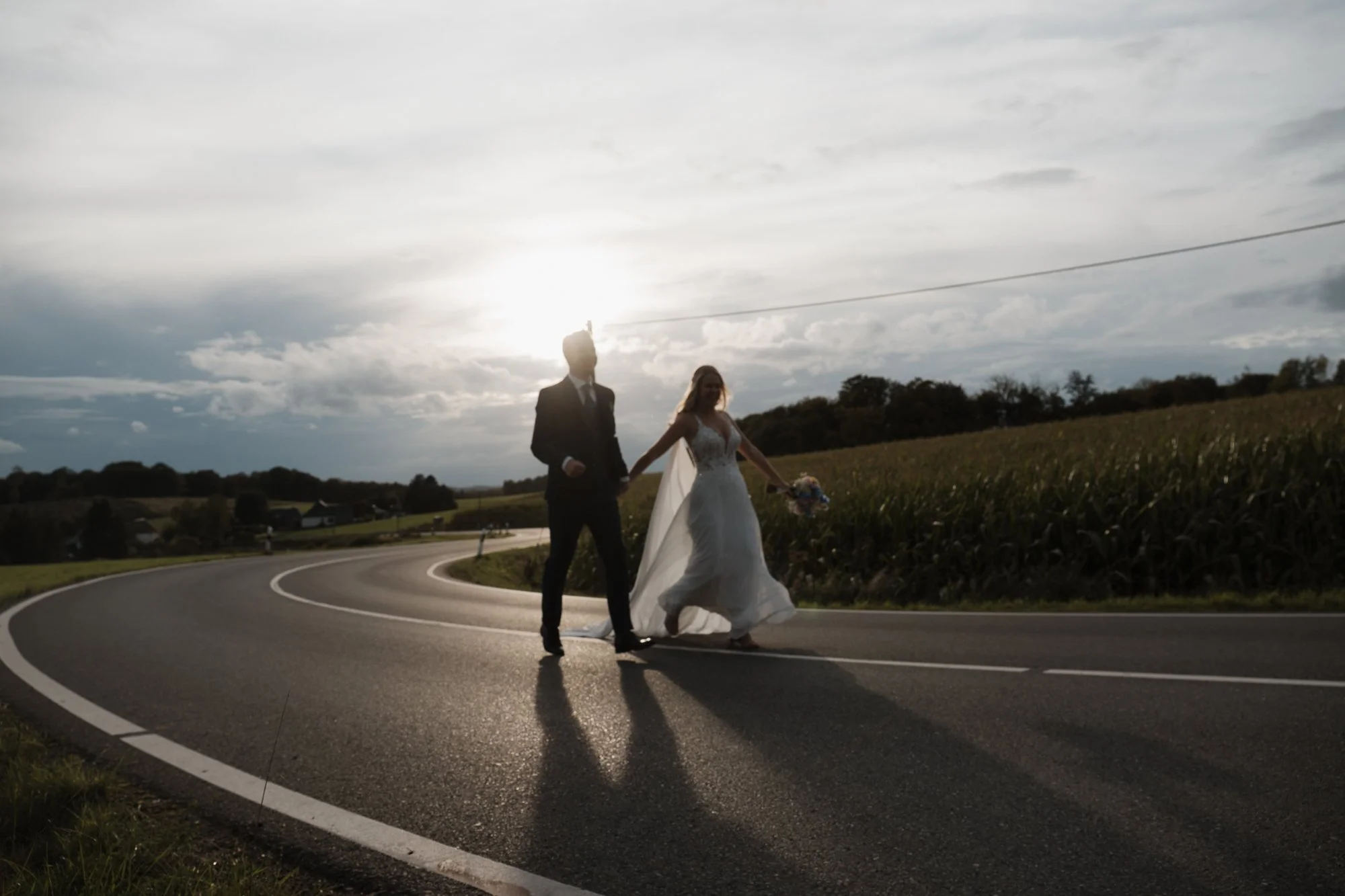 Ein Brautpaar hält Händchen auf einer kurvigen Landstraße bei Sonnenuntergang, umgeben von Feldern.