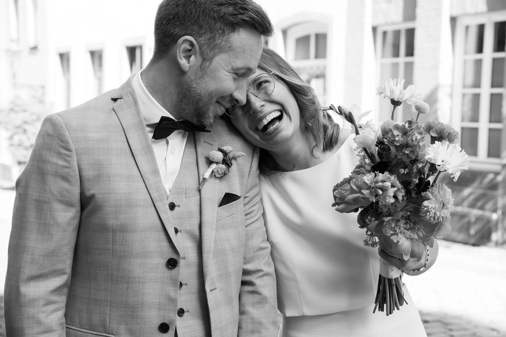 Ein lachendes Paar, wahrscheinlich Braut und Bräutigam, bei ihrer Hochzeit, gemeinsam lachend und umarmend, mit Blumen, im Freien vor einem Gebäude.