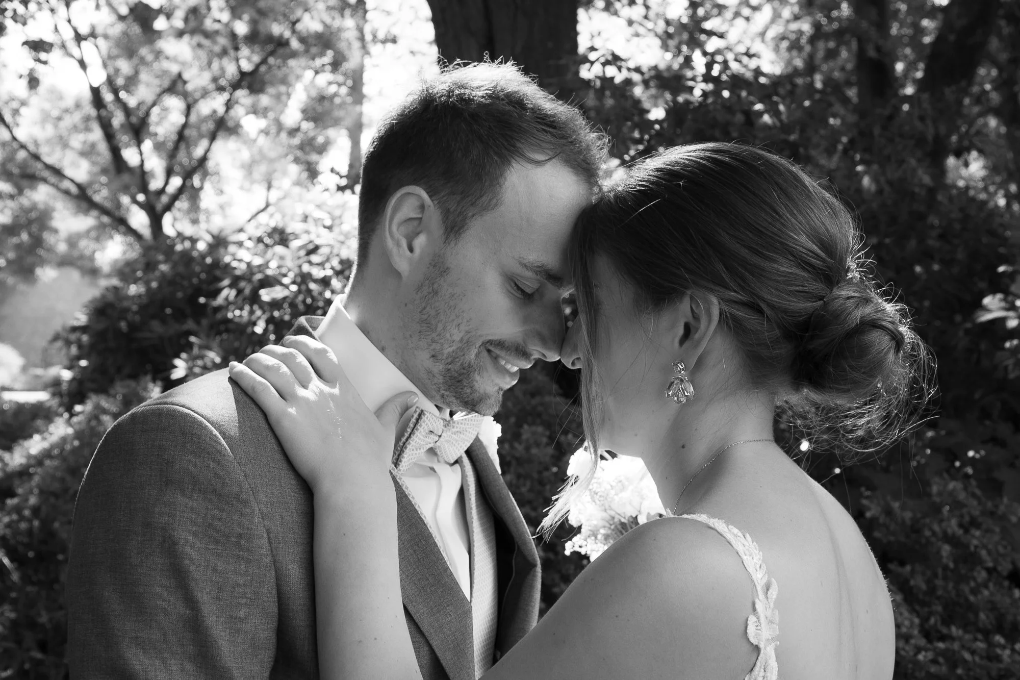 Ein Paar bei einer Hochzeit stehend mit Stirn an Stirn im Freien, im Hintergrund Bäume, schwarz-weiße Fotografie.