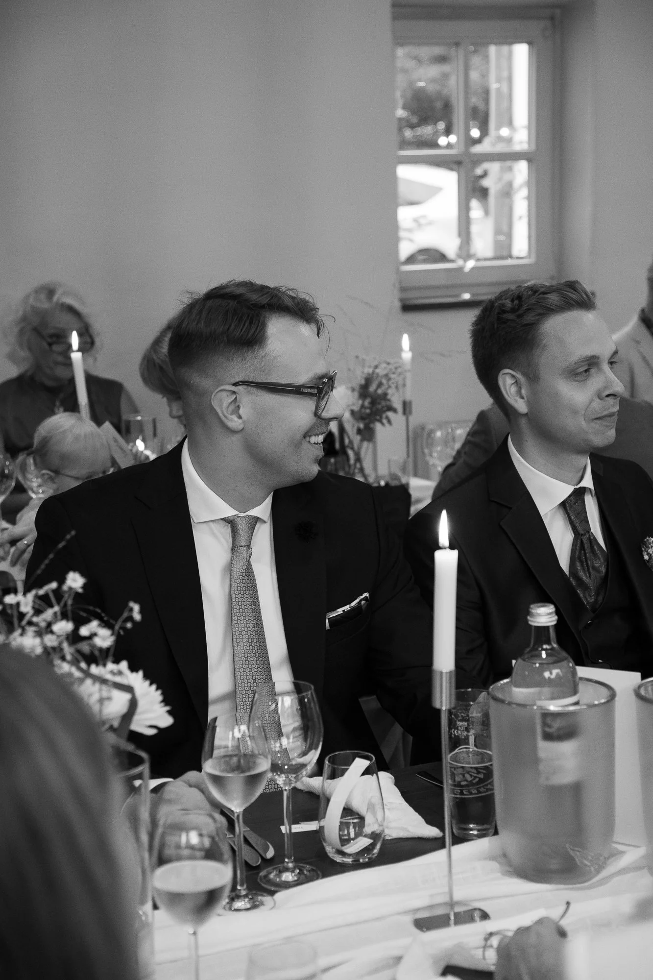 Zwei Männer in Anzügen, die an einem Essensabend sitzen, bei dem Kerzenlicht und Wein. Im Hintergrund sind weitere Gäste und eine helle Wand mit Fenster.