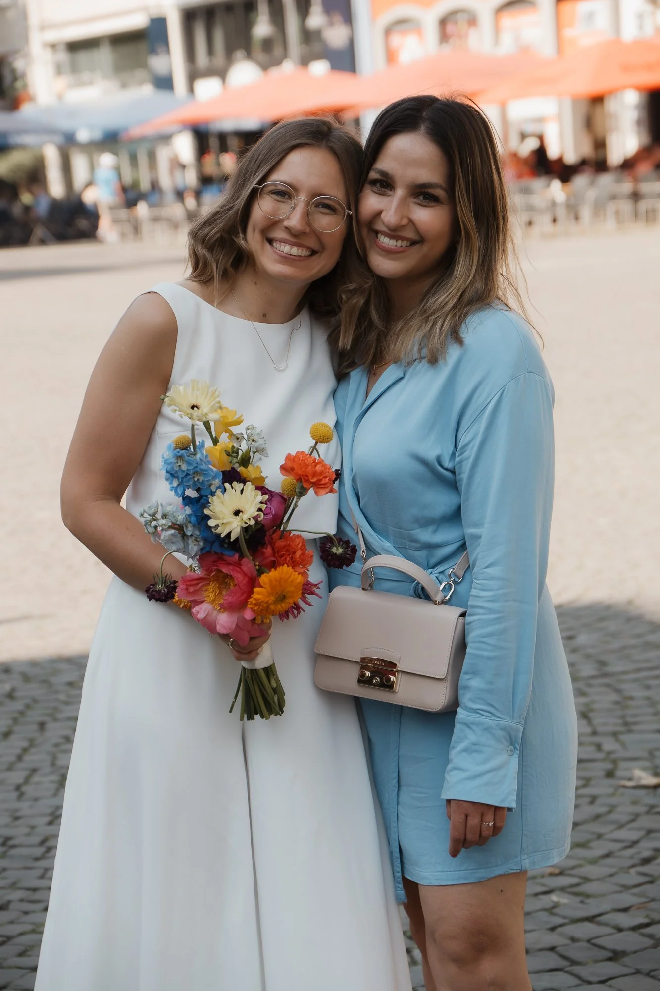 Zwei lachende Frauen stehen gemeinsam draußen, eine trägt ein weißes Kleid und hält einen bunten Blumenstrauß, die andere trägt ein hellblaues Kleid mit einer hellen Handtasche.