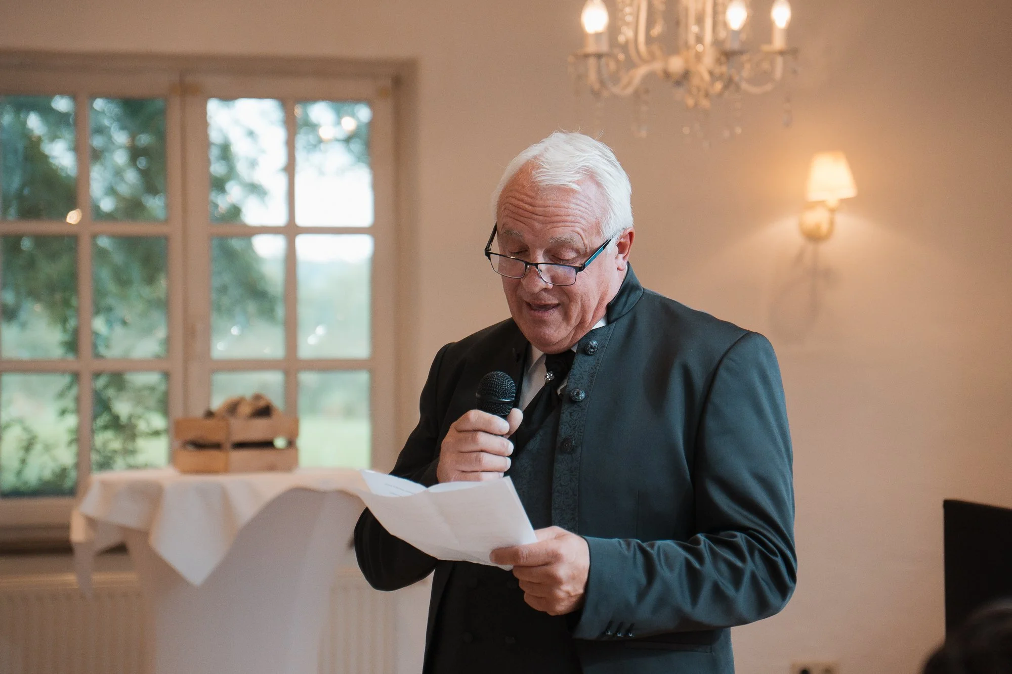 Ein älterer Mann mit weißen Haaren und Brille, trägt einen schwarzen Anzug, hält ein Mikrofon in der einen Hand und liest aus einem Papier in der anderen Hand vor. Im Hintergrund sind große Fenster mit Blick ins Grüne, eine Stehlampe und ein Kronleuc
