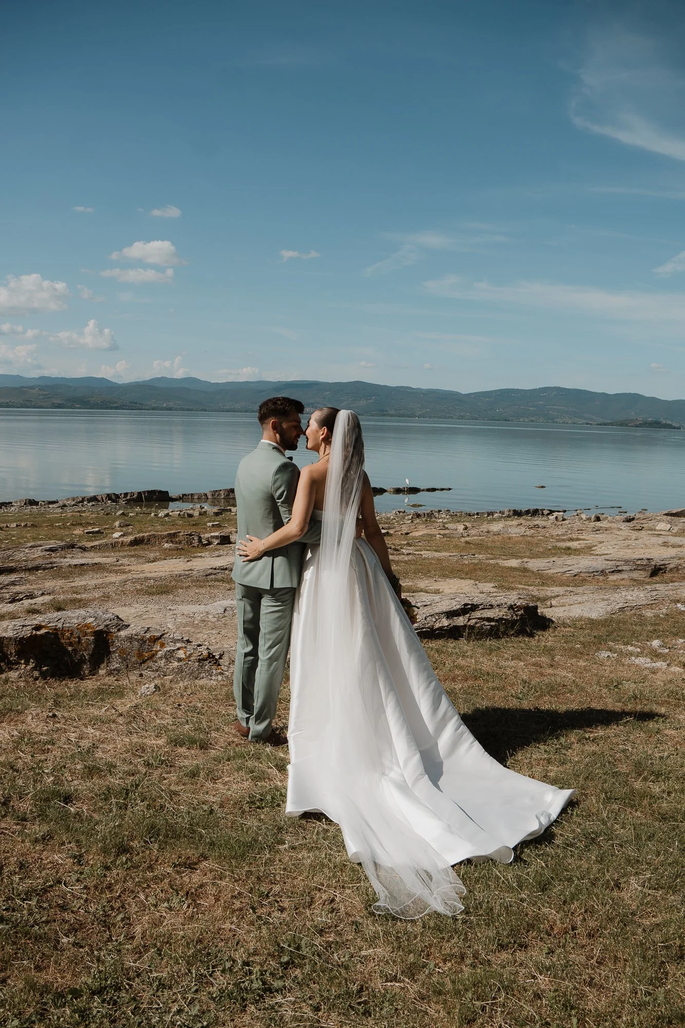 Ein frisch vermähltes Paar steht außen an einem See, umgeben von Bergen, trägt Hochzeitskleidung, lächelt sich an.