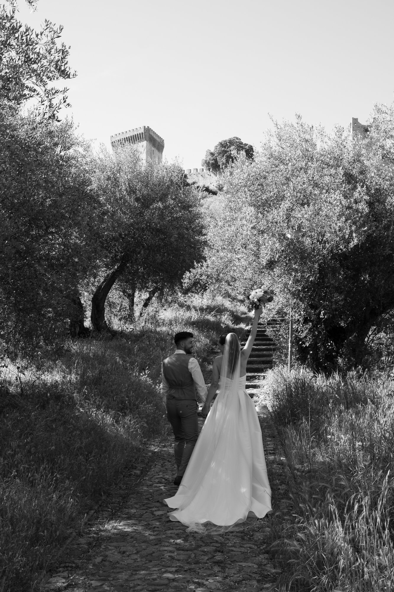 Ein Brautpaar läuft auf einem Naturweg, die Braut hält einen Blumenstrauß hoch, im Hintergrund Bäume und eine Burg