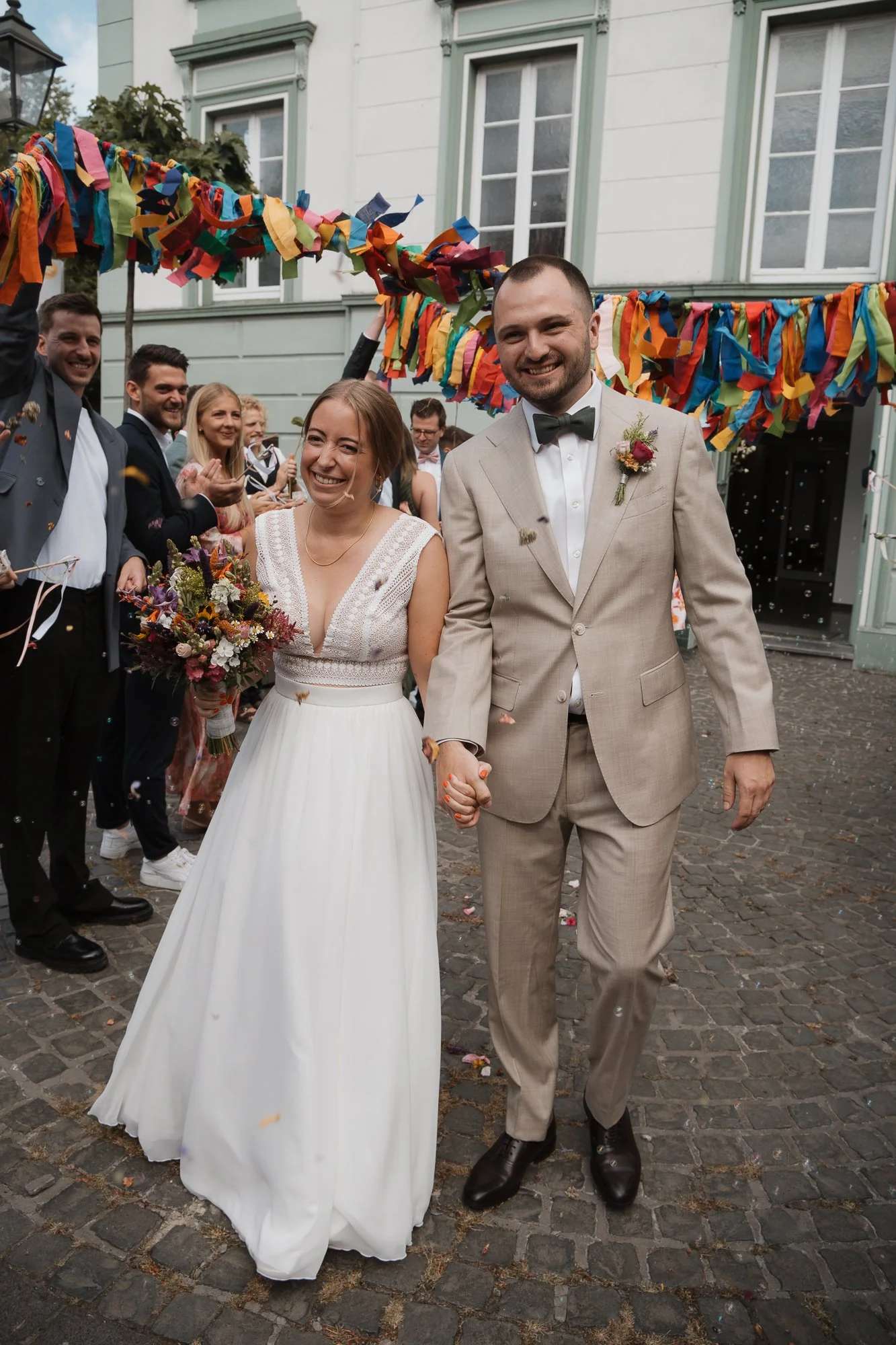 Ein glückliches Brautpaar bei ihrer Hochzeit, umgeben von Freunden, mit bunten Bändern und Konfetti im Hintergrund.