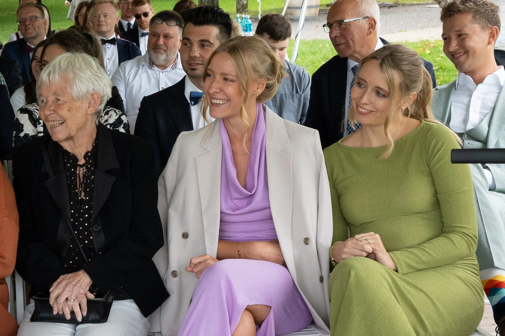 Gruppen von Menschen, die bei einer Veranstaltung im Freien sitzen, lachen und sich unterhalten. Im Vordergrund drei Frauen, eine im schwarzen Kleid, eine in weißem Mantel mit lila Kleid und eine in grüner Kleidung, die kämpfen.