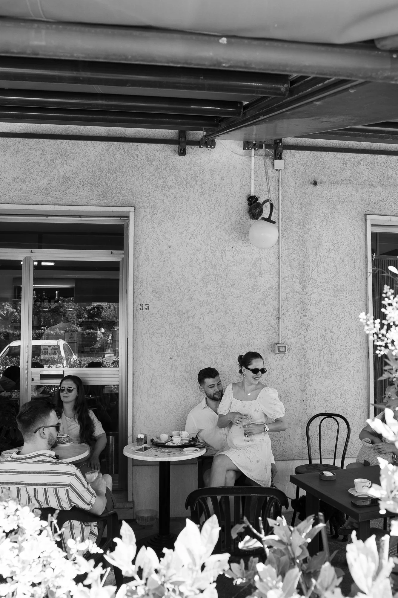 Menschen sitzen auf einer Terrasse, trinken Kaffee und unterhalten sich. Im Vordergrund sind Blätter und Blüten, im Hintergrund eine Wand mit Fenstern und einer Außenlampe.