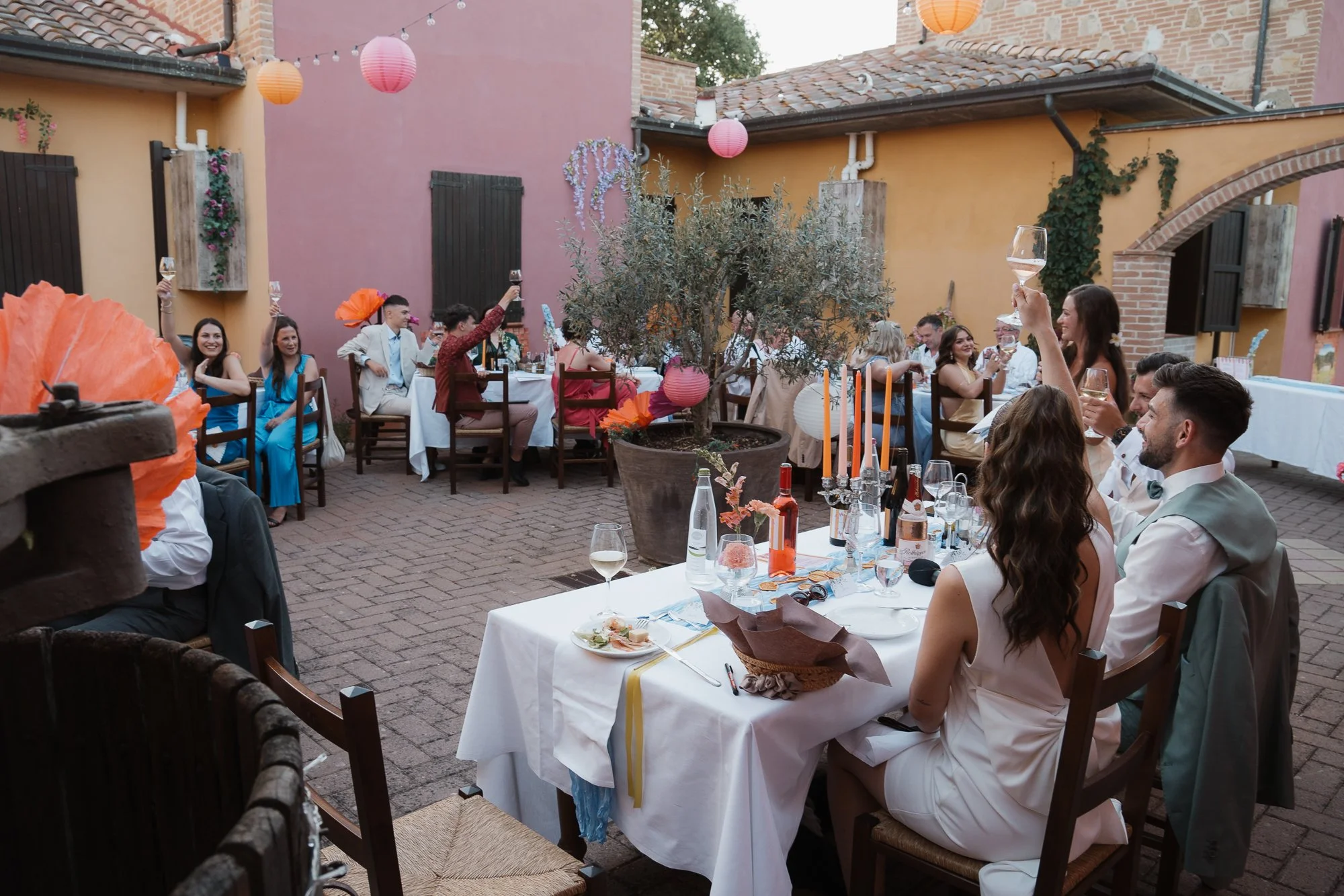 Junge Menschen feiern eine Gartenparty oder Hochzeit im Freien, heben Gläser und haben eine festliche Stimmung. Es gibt Tische mit Kerzen, Wein und Essen, bunte Laternen und Dekorationen im Innenhof eines Gebäudes mit gelben und pinkfarbenen Wänden.