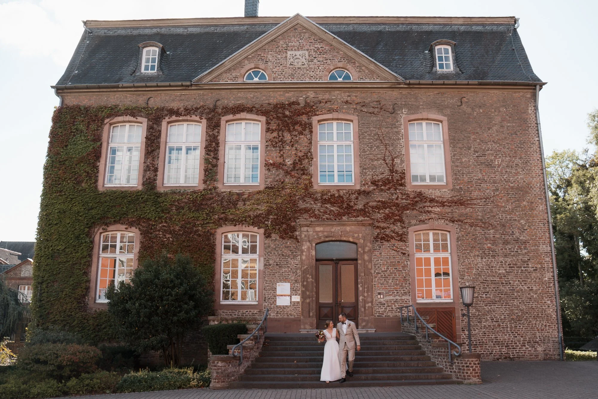 Ein Brautpaar in Hochzeitskleidung auf den Stufen eines alten Backsteingebäudes, umgeben von Bäumen und Pflanzen.