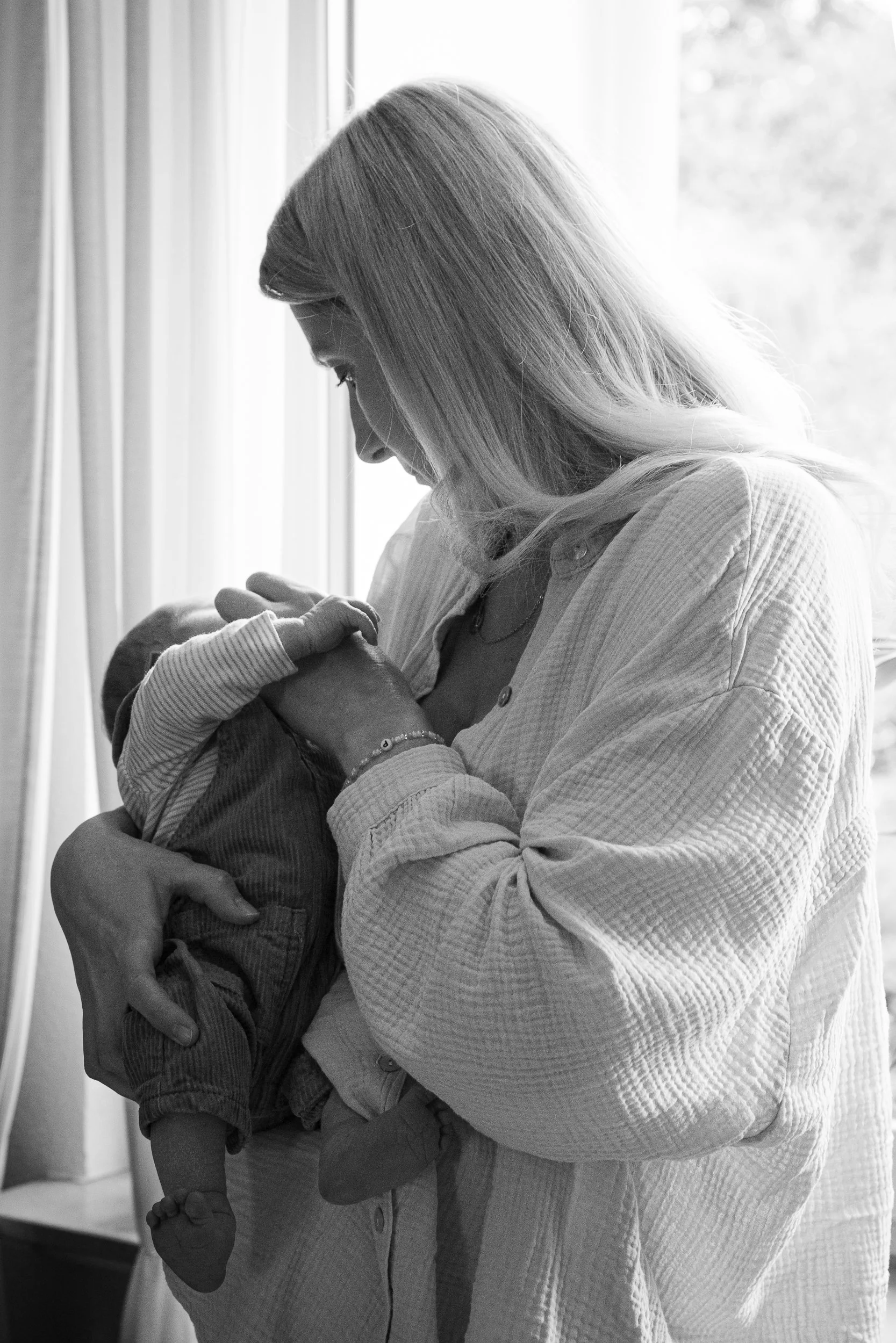 Eine Frau hält ein Baby im Arm und küsst es zärtlich, während sie an einem Fenster steht.