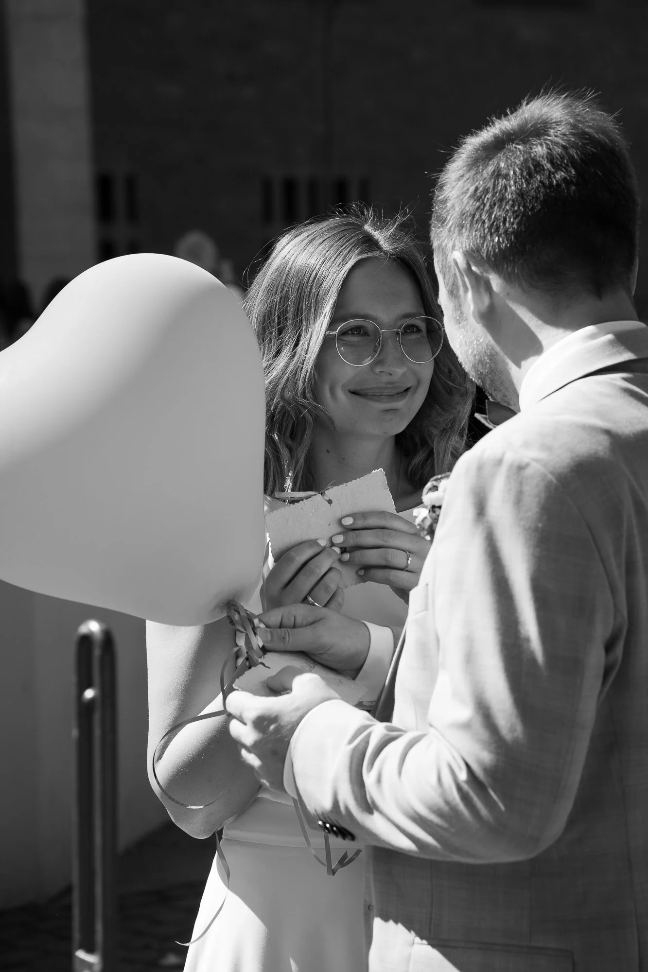 Ein Brautpaar bei der Hochzeitszeremonie, die Frau trägt eine Brille und hält einen Brief, während der Mann ein Herzballon hält.