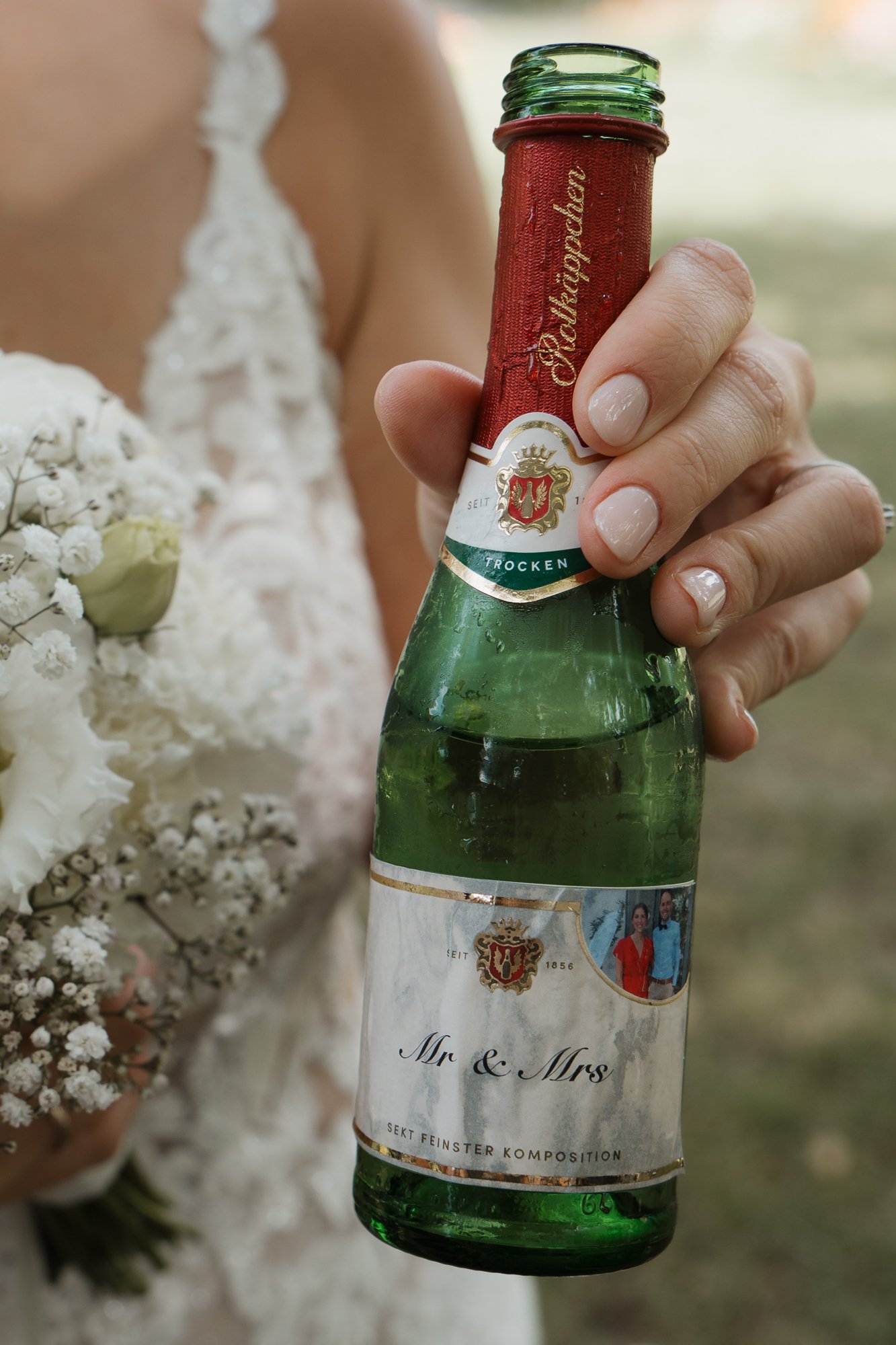 Eine Hand hält eine Flasche Sekt mit einem Etikett, auf dem 'Mr & Mrs' steht, neben einer Braut mit Hochzeitsstrauß.