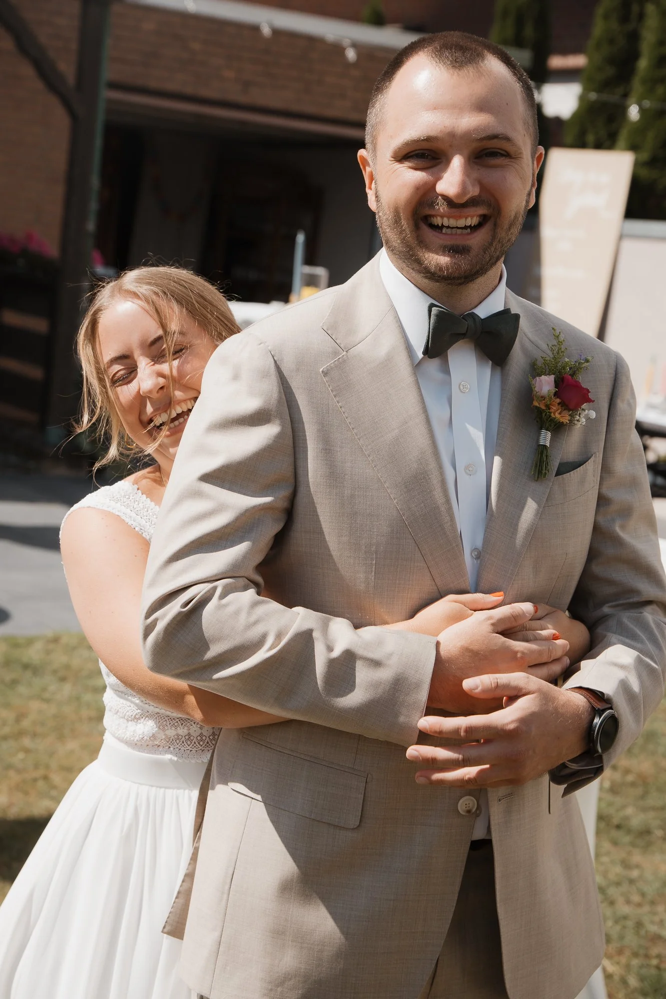 Ein lachendes Brautpaar beim Hochzeitsshooting im Freien, die Braut umarmt den bräutigen Rücken.