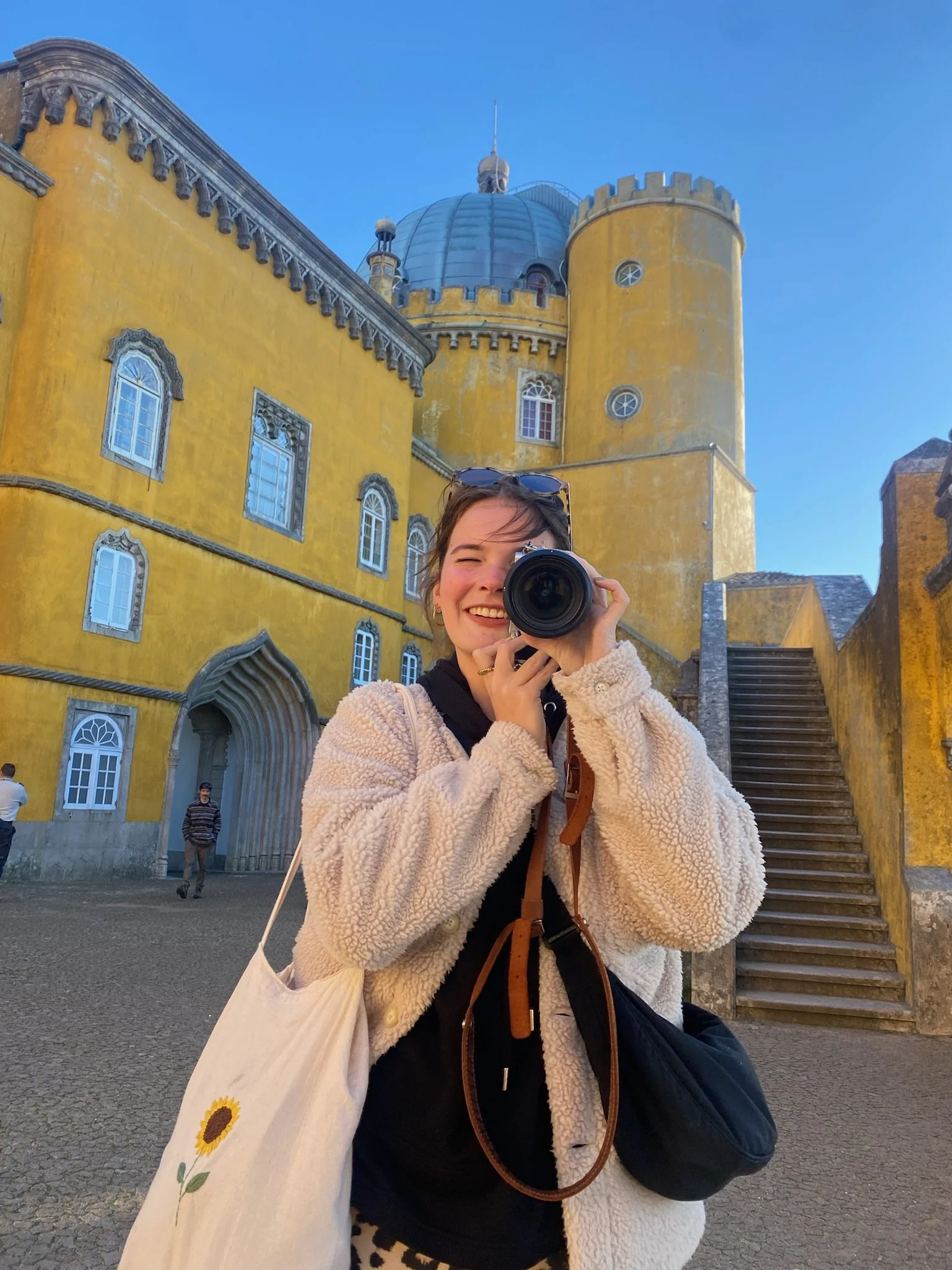 Junge Frau fotografiert mit Kamera vor einem gelben Schloss mit runden Türmen unter klarem blauen Himmel.