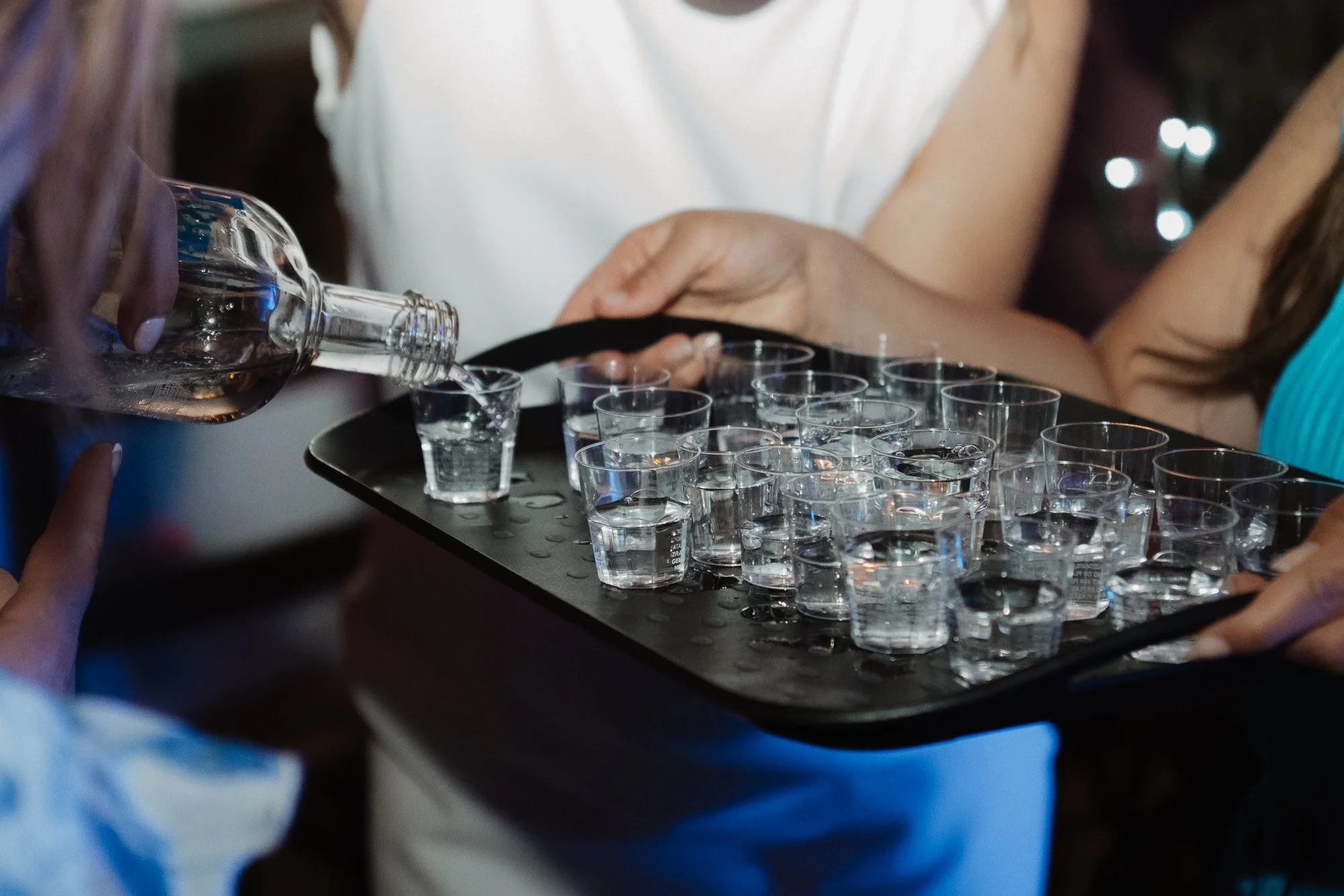 Person gießt alkoholisches Getränk in kleine Schnapsgläser auf einem Tablett bei einer Party.