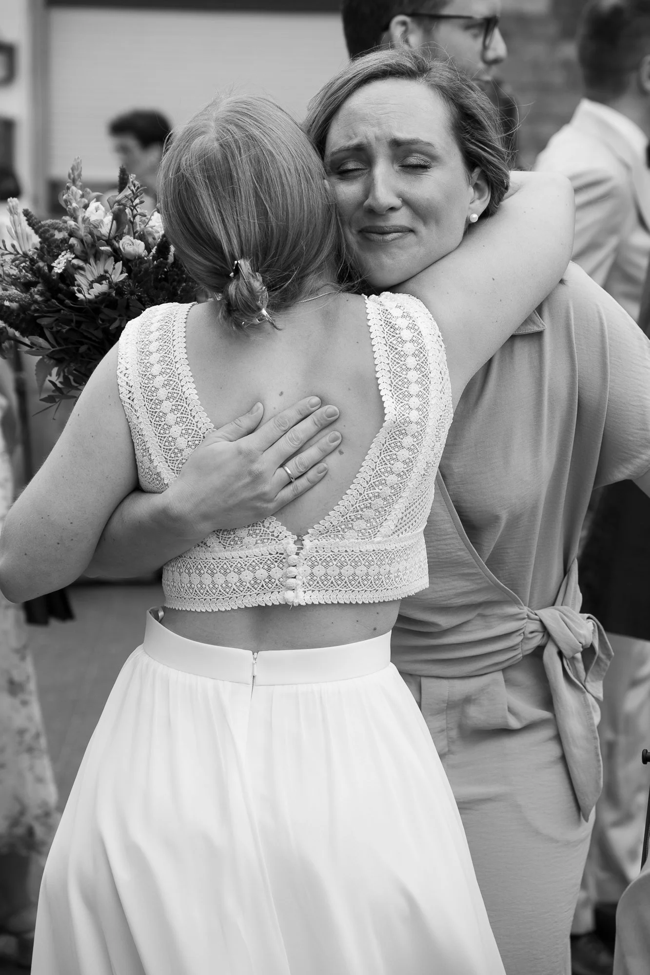 Zwei Frauen umarmen sich, eine Frau mit einem weißen Kleid und einer Lace-Top, die andere mit kurzem Haar und einer Bluse, bei einer emotionalen Szene im Freien.