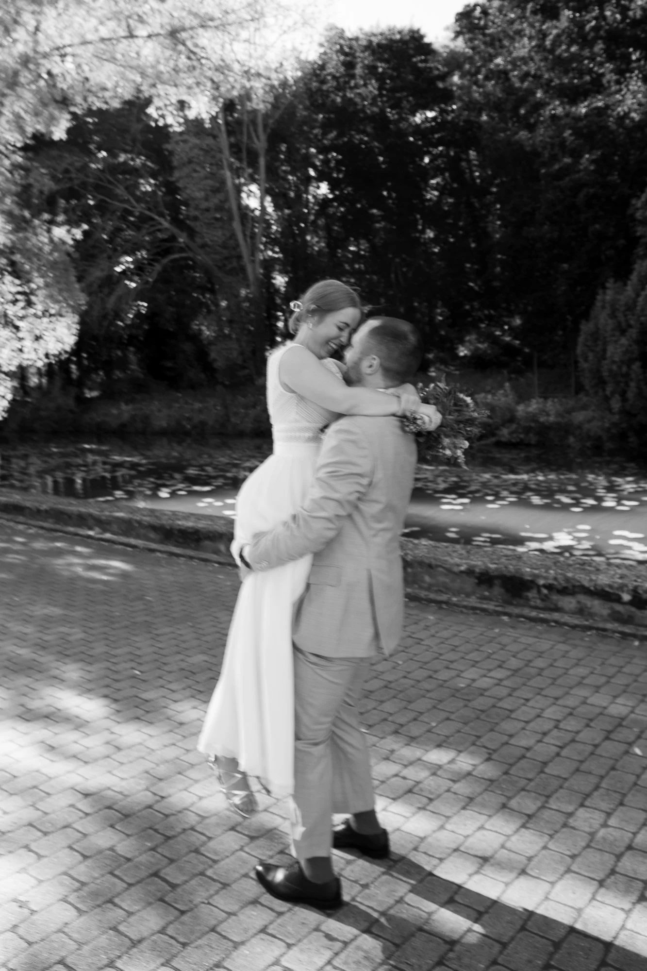 Ein Mann hebt eine Frau in einem weißen Kleid hoch, beide lächeln und umarmen sich liebevoll, im Freien bei Tageslicht, schwarz-weiß Fotografie.