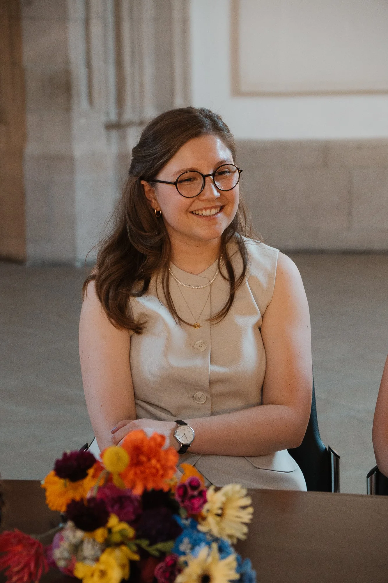 Eine lächelnde Frau mit Brille und langem braunem Haar sitzt an einem Tisch mit bunten Blumen. Sie trägt eine cremefarbene ärmellose Bluse, eine Uhr, Halsketten und Ohrringe. Das Ambiente wirkt wie ein eleganter Raum mit Steinwänden.