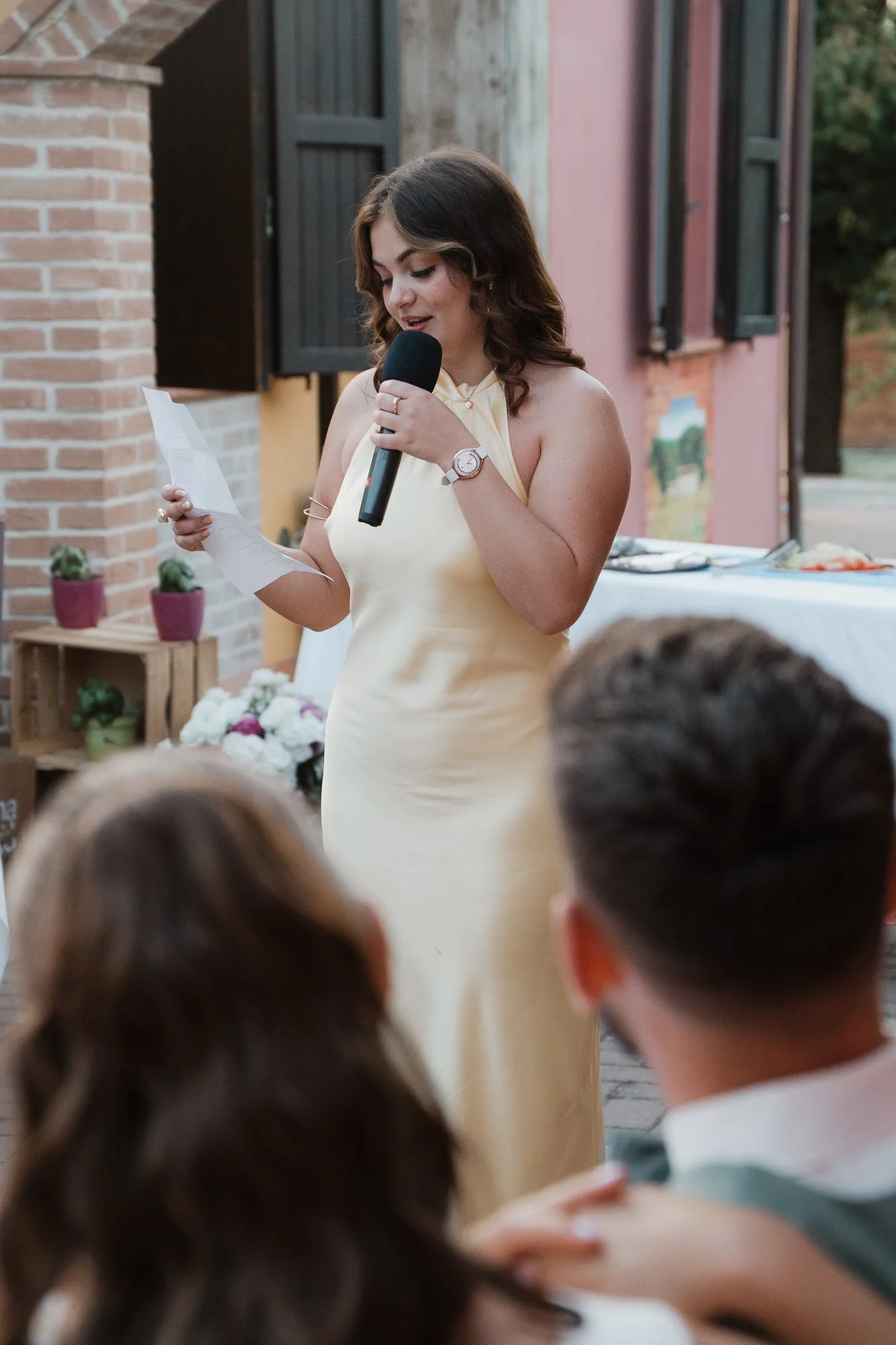 Eine junge Frau in einem cremefarbenen Kleid hält ein Mikrofon in der Hand und liest aus einem Papier während einer Veranstaltung im Freien. Im Vordergrund sind die Köpfe zweier Personen sichtbar, die auf die Frau schauen.