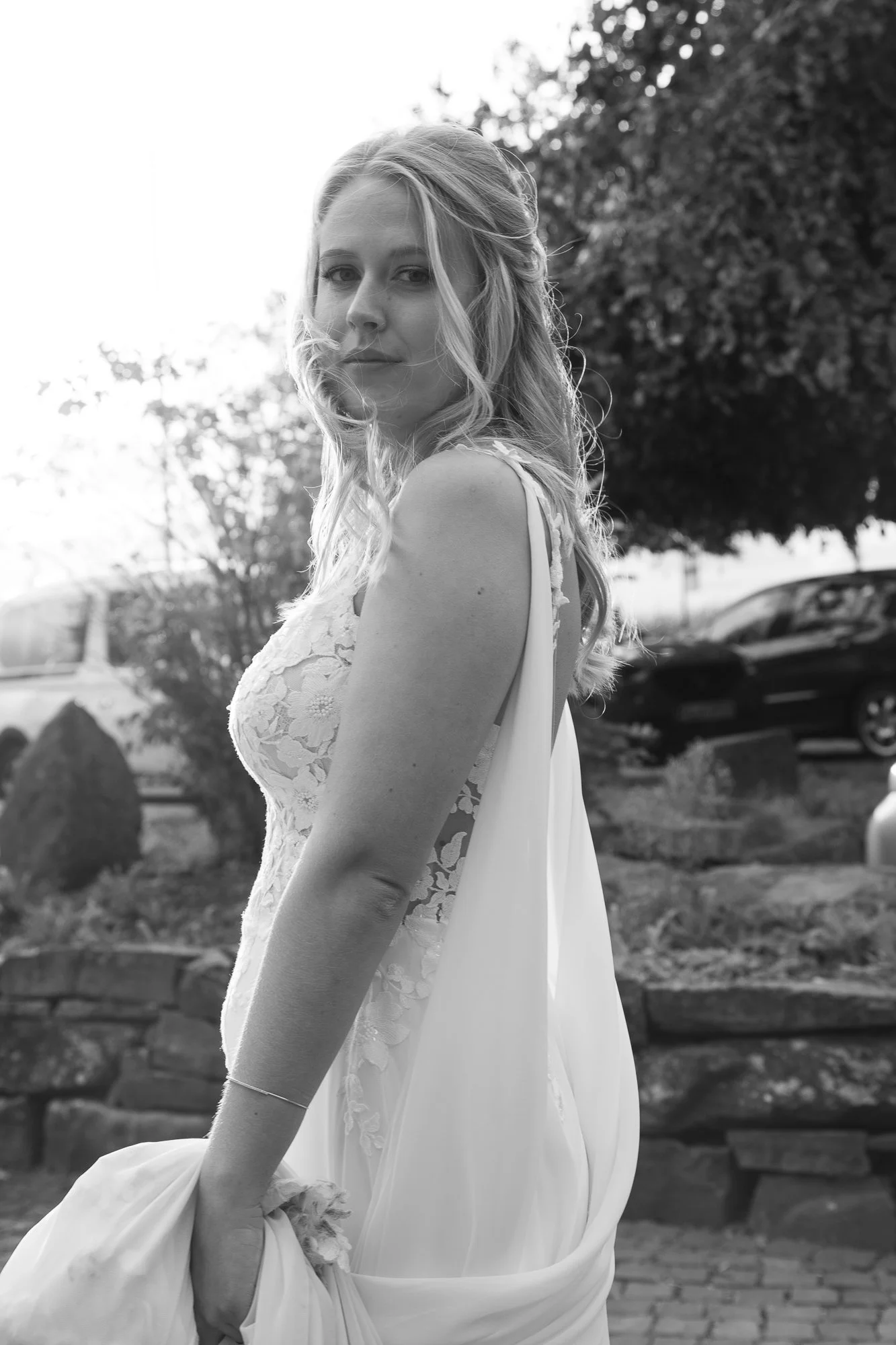 Schwarz-weiß-Foto eines jungen Mädchens in einem Brautkleid, das in einem Garten steht, mit Bäumen und Autos im Hintergrund.