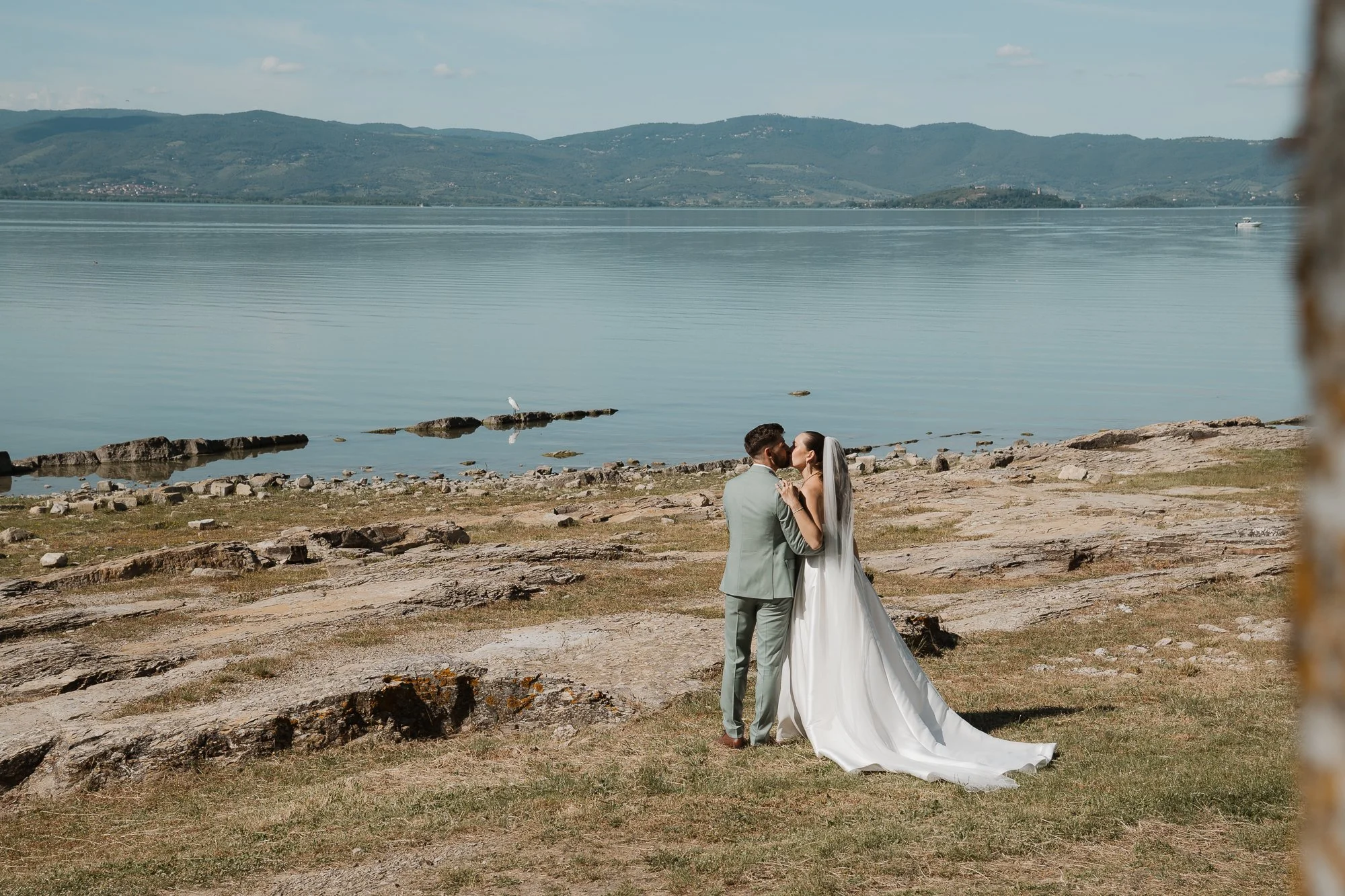 Ein Brautpaar in Hochzeitskleidung steht am Ufer eines Sees und küsst sich bei schönem Wetter.