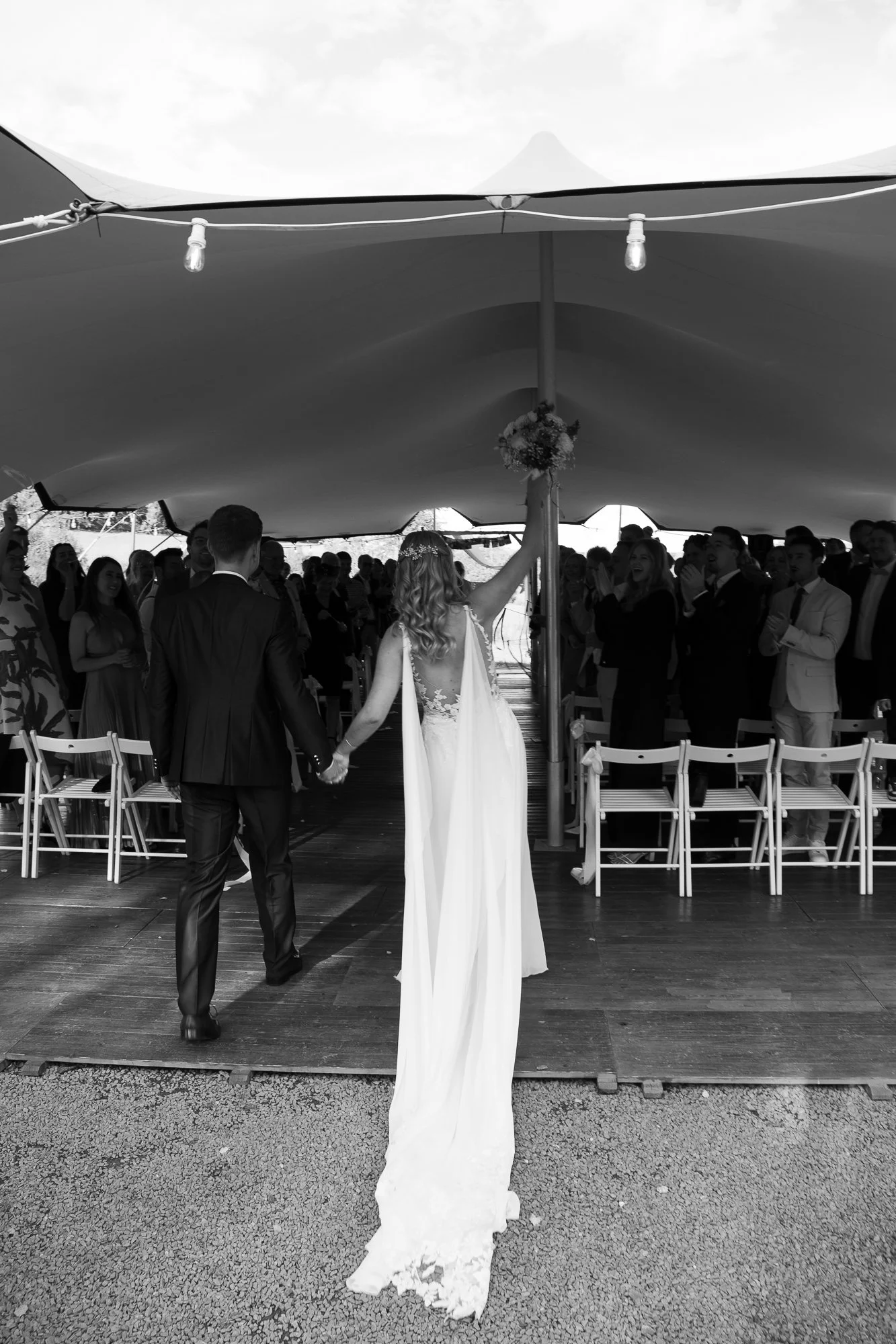 Braut und Bräutigam bei ihrer Hochzeitszeremonie in einem Zelt, die Braut hält einen Blumenstrauß hoch, die Gäste stehen und sitzen und beobachten die Zeremonie.