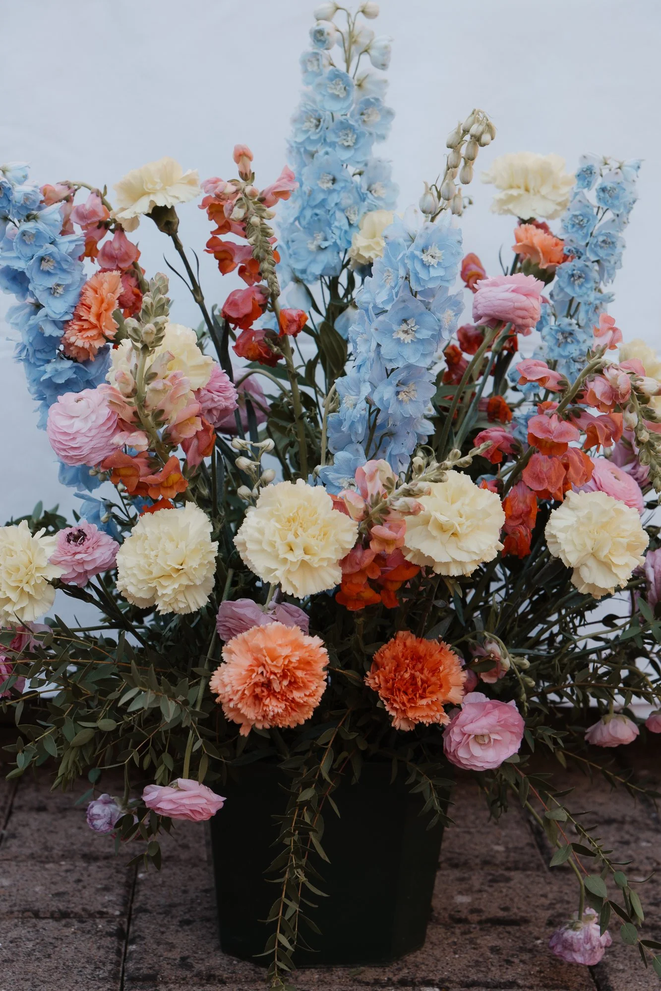 Bunte Blumen in einer schwarzen Vase, darunter zweigen sich grüne Blätter ab.