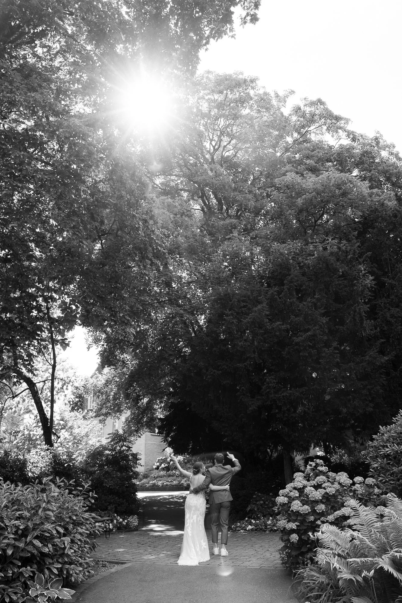 Ein Brautpaar geht durch einen Garten, umgeben von Bäumen und Blumen. Es ist sonnig, und die Sonne scheint durch die Blätter.