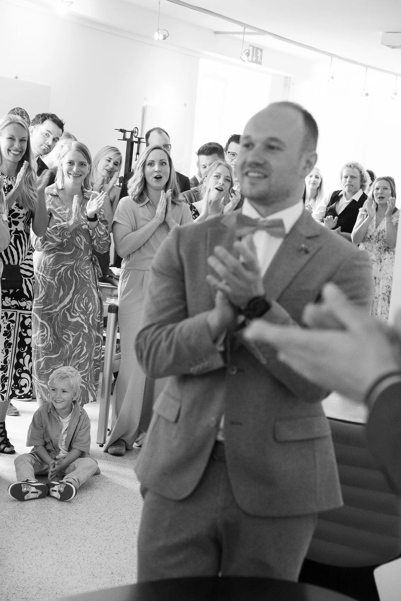 Ein Mann in einem Anzug und Fliege wird in einem Raum von einer Gruppe fröhlicher und überraschter Menschen umgeben, während ein kleines Kind auf dem Boden sitzt und zuschaut.