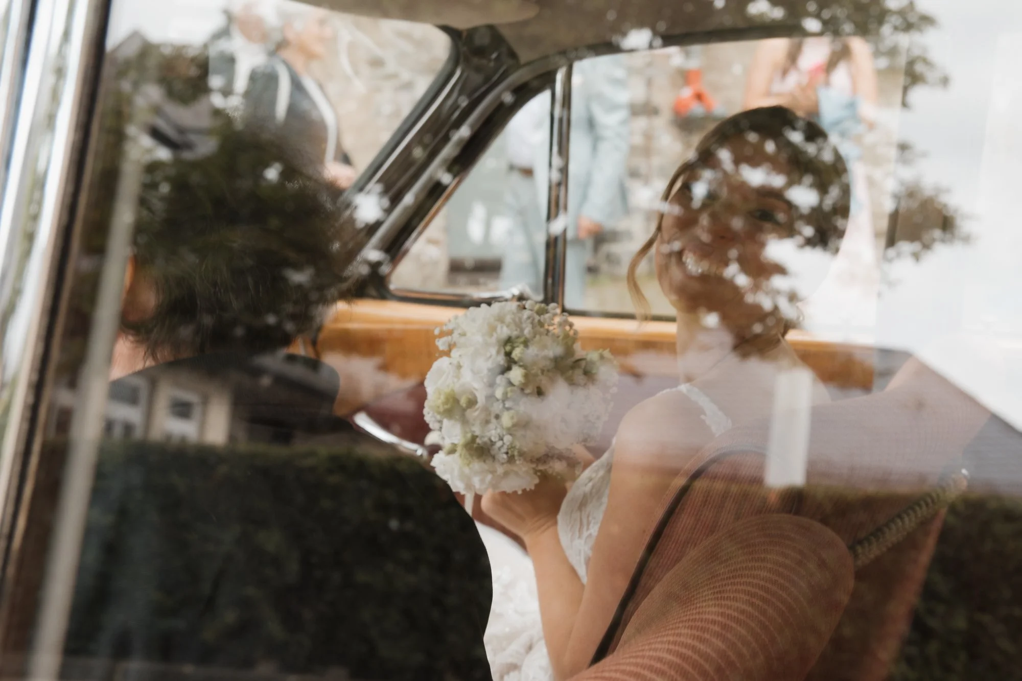 Braut sitzt in einem Oldtimer, blickt nach vorne und hält einen Blumenstrauß.