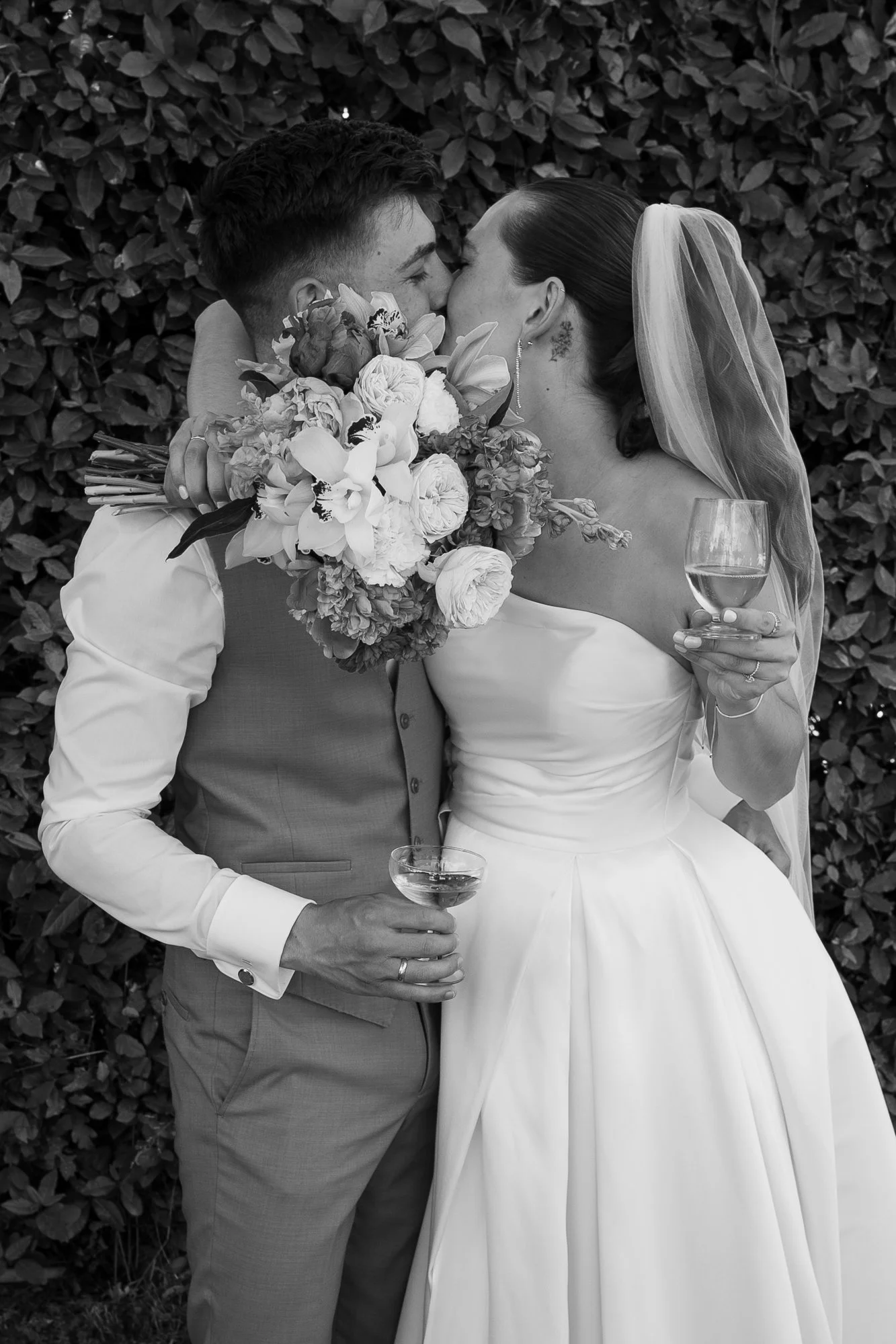 Braut und Bräutigam küssen sich bei ihrer Hochzeit, während sie mit Sekt anstoßen, im Hintergrund eine Hecke.