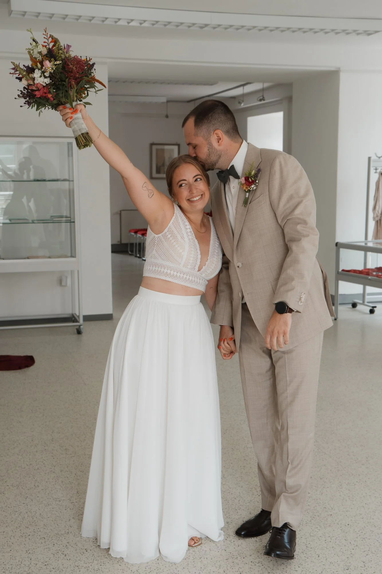 Ein glückliches Brautpaar bei ihrer Hochzeit, die Braut hält einen Blumenstrauß hoch, der Bräutigam küsst die Braut auf die Stirn