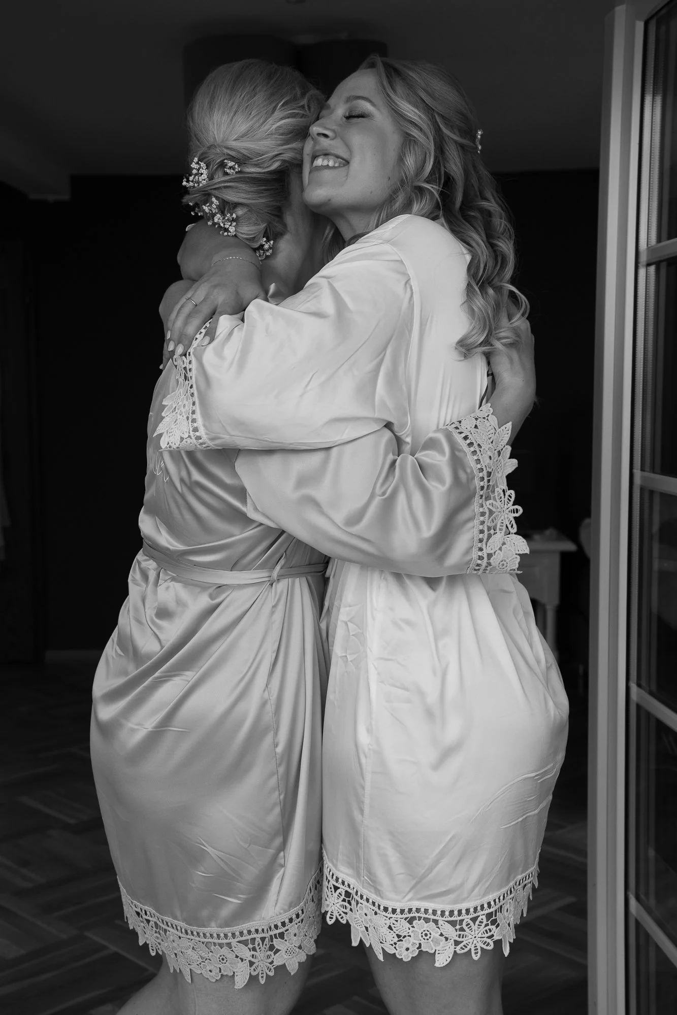 Zwei Frauen in Seidenbademänteln, die sich umarmen und lachen, im Badezimmer, schwarz-weiß Foto.