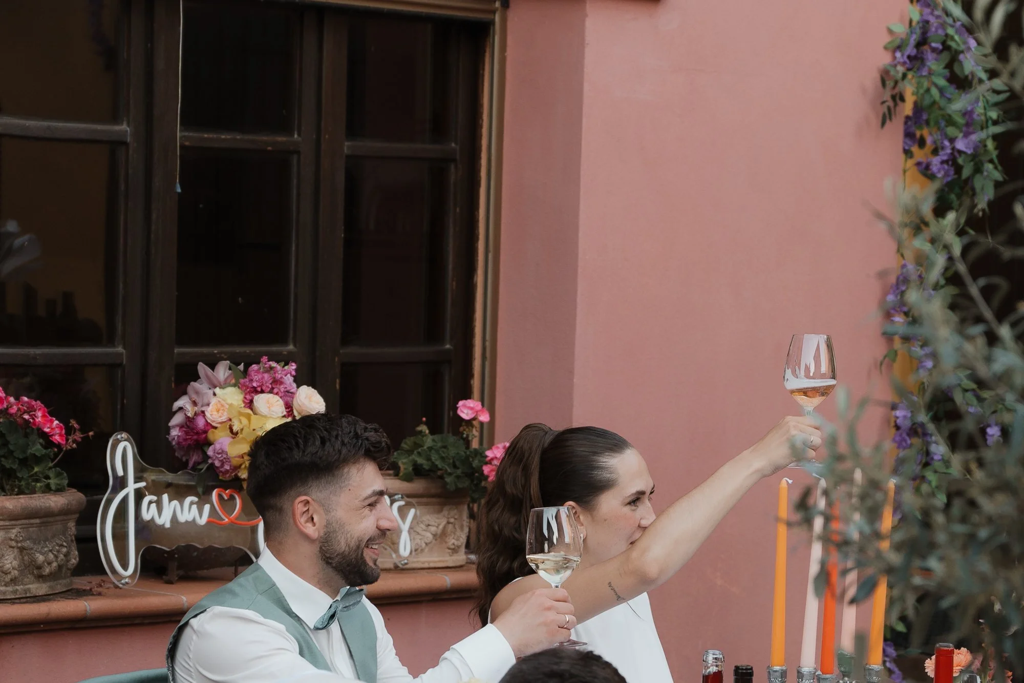 Ein lachendes Paar bei einer Feier, beide halten Weingläser, die Frau hebt ihr Glas in die Luft, im Hintergrund sind Blumen und eine rosa Wand.