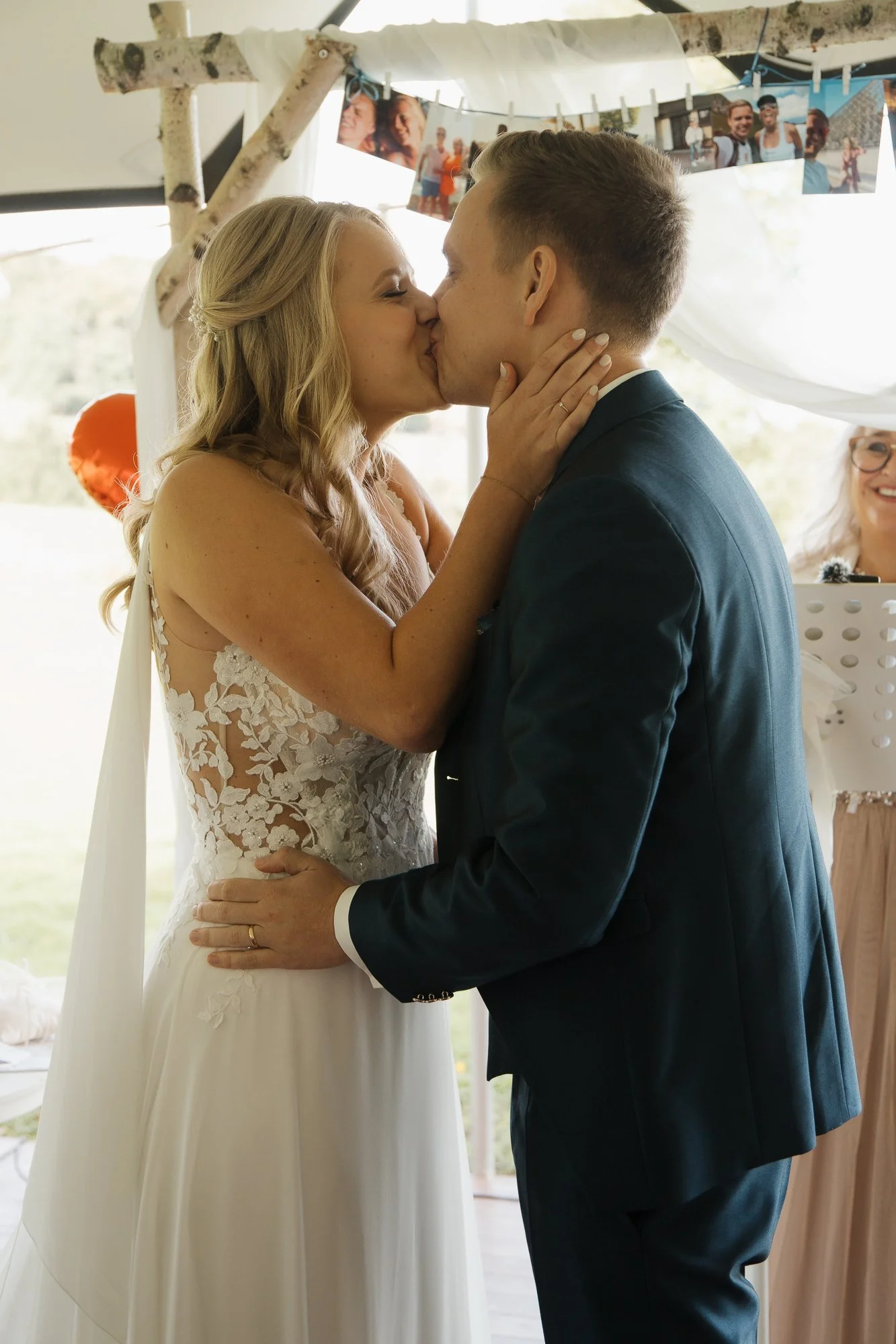 Ein Brautpaar beim Kuss während ihrer Hochzeitsfeier, im Hintergrund eine Fotogirlande. Die Frau trägt ein weißes Hochzeitskleid mit Spitze, der Mann einen dunklen Anzug.