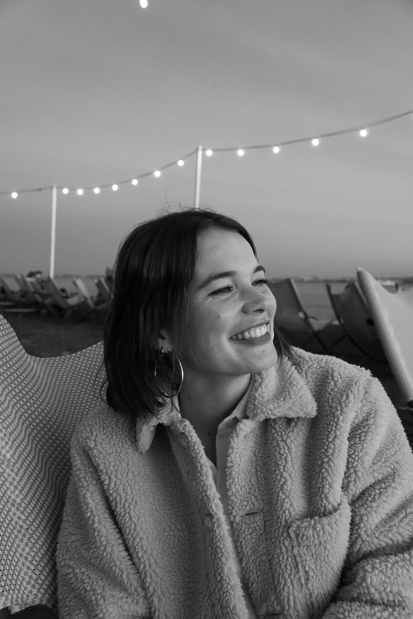 Junge Frau sitzt bei Sonnenuntergang am Strand, trägt eine Fleece-Jacke und lächelt.