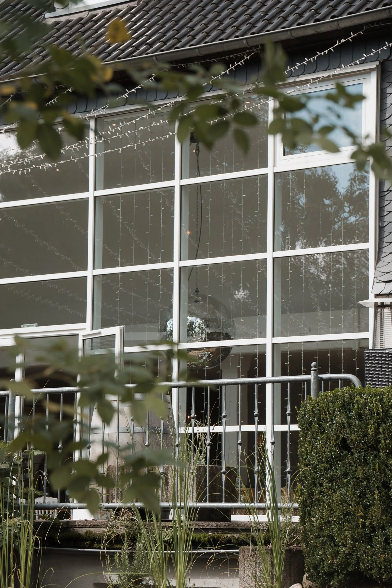 Großer Fensterbereich eines Hauses mit mehreren quadratischen Fenstern, Dekorative Lichterketten, Büsche und Pflanzen im Vordergrund, Dachziegel sichtbar.