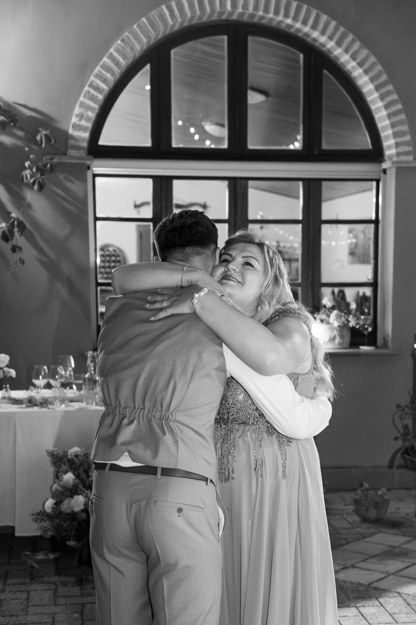 Ein Mann und eine Frau umarmen sich in einem festlichen Raum mit einer großen Fensterfront, Dekorationen und Tischen im Hintergrund, schwarz-weiß