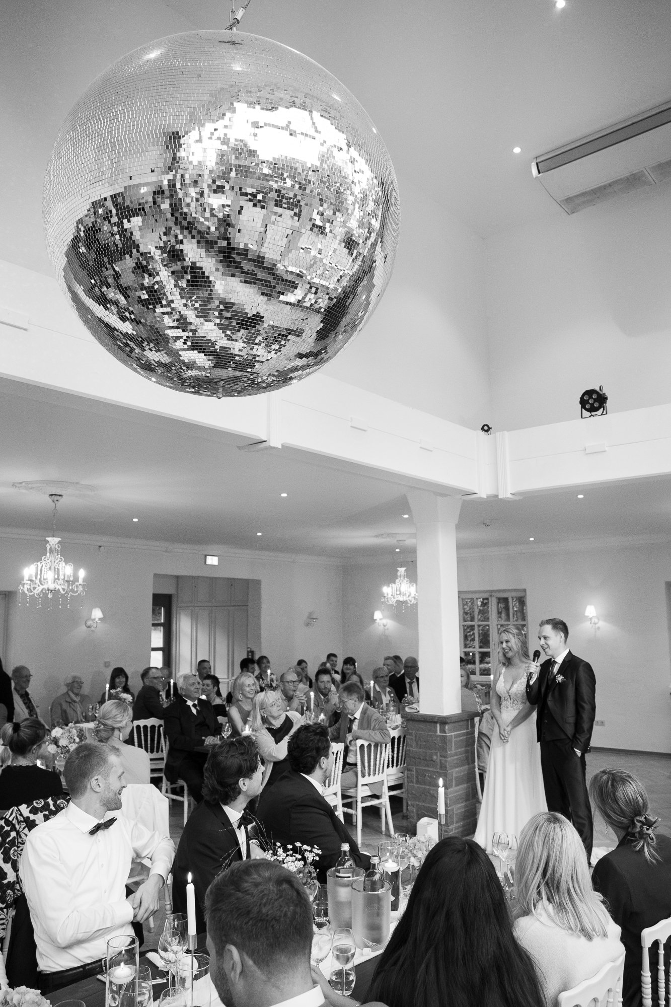 Ein Hochzeitsfest in einem festlich geschmückten Saal mit einem Disco-Kugel-Hängel am Decke. Zwei Menschen, vermutlich das Brautpaar, sprechen vor einer applaudierenden Gästegruppe. Es gibt Kerzen, Blumen und Tische mit Getränken.