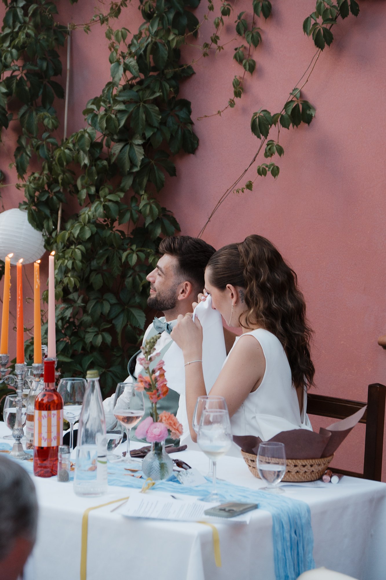 Ein Hochzeitspaar sitzt an einem festlich gedeckten Tisch bei ihrer Feier, der Mann trägt einen Anzug mit Fliege, die Frau eine weiße Kleidung, im Hintergrund eine rosa Wand mit grünen Kletterpflanzen.